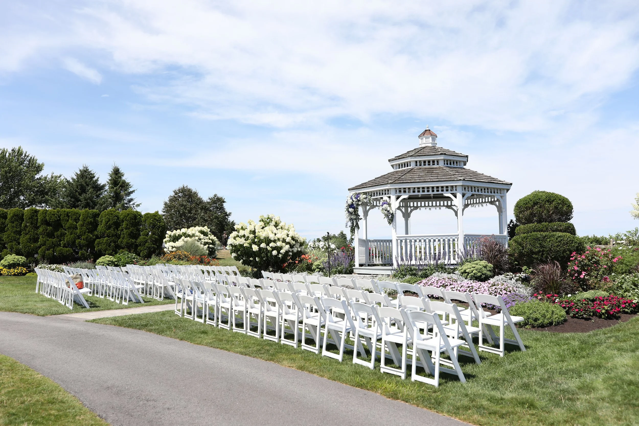 Outdoor wedding setup with white chairs arranged in rows in front of a white gazebo, surrounded by colorful flowers and greenery under a partly cloudy sky.
