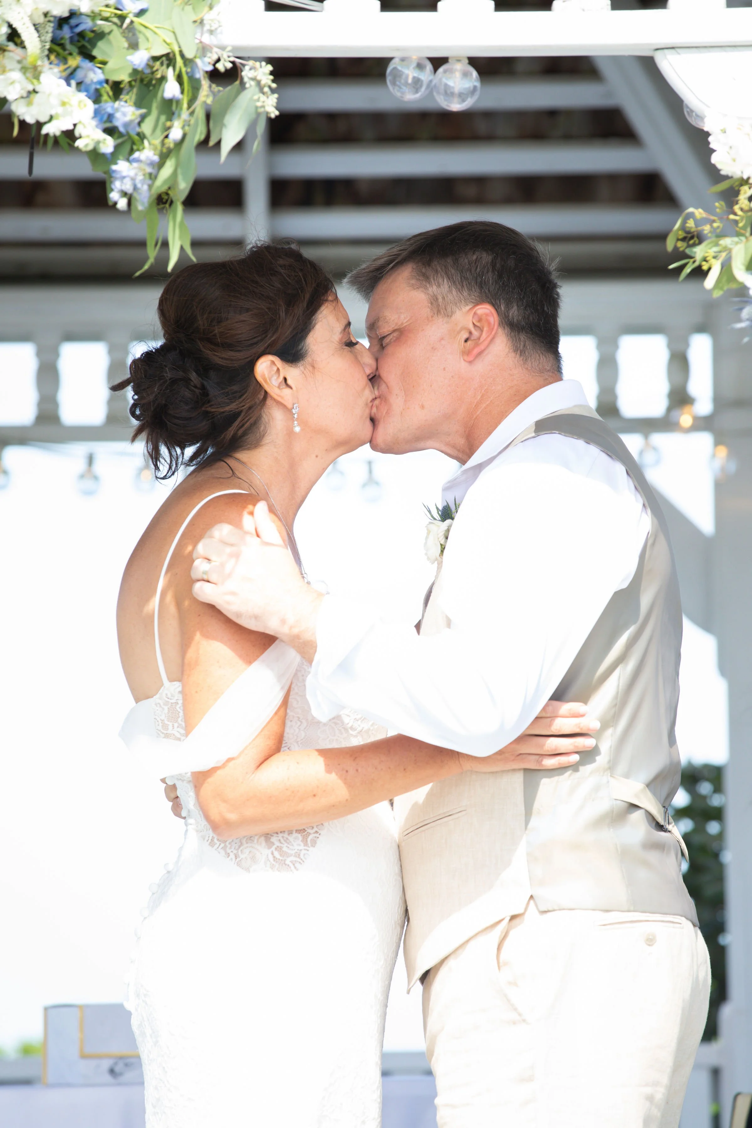 A bride and groom share a kiss during a wedding ceremony outdoors in bright sunlight, with floral decorations and a white wooden structure in the background.