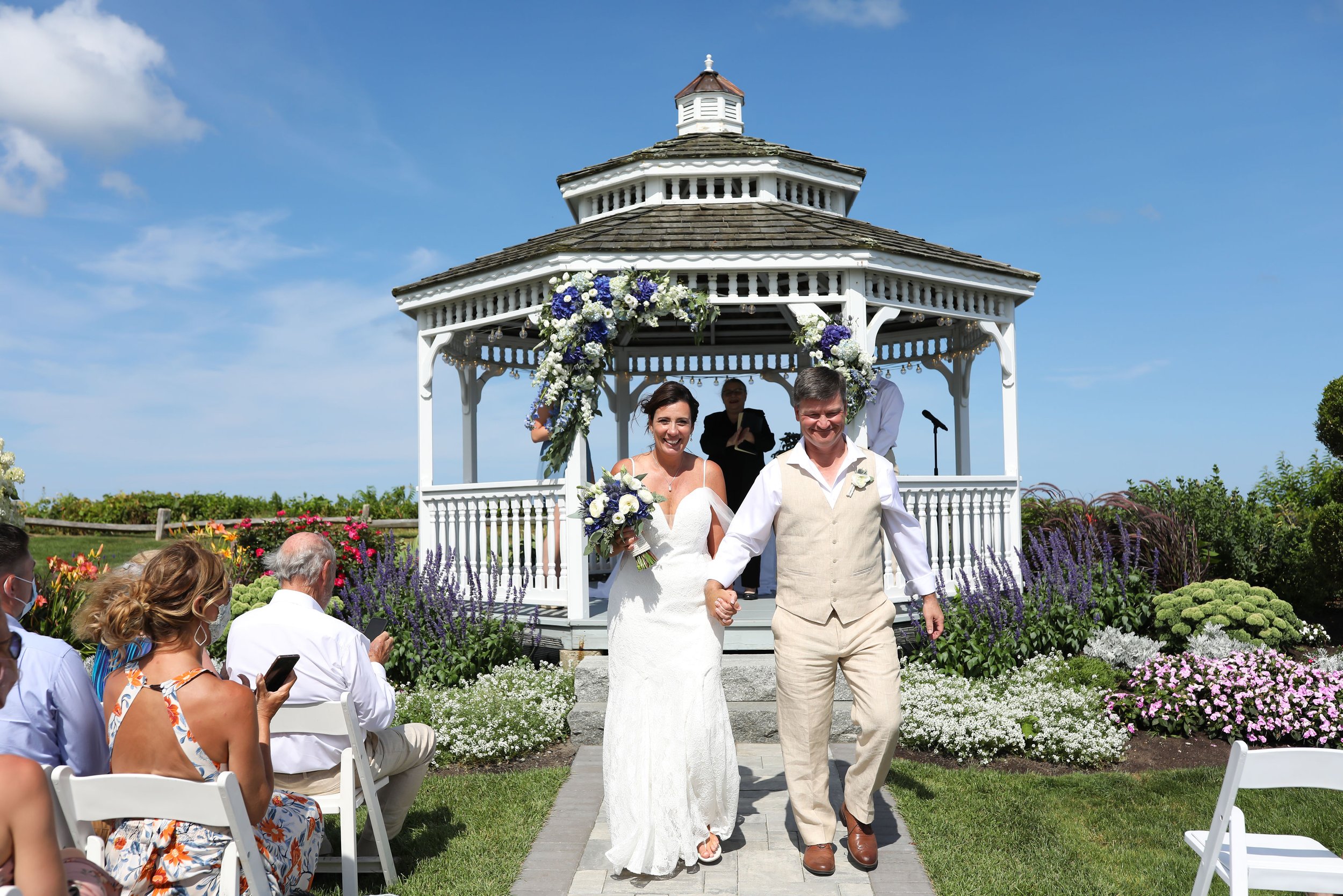 A wedding couple walking hand-in-hand down an outdoor garden aisle, with the bride in a white dress holding a bouquet and the groom in a beige suit. They are smiling as guests seated on white chairs watch them, and a church or gazebo decorated with f