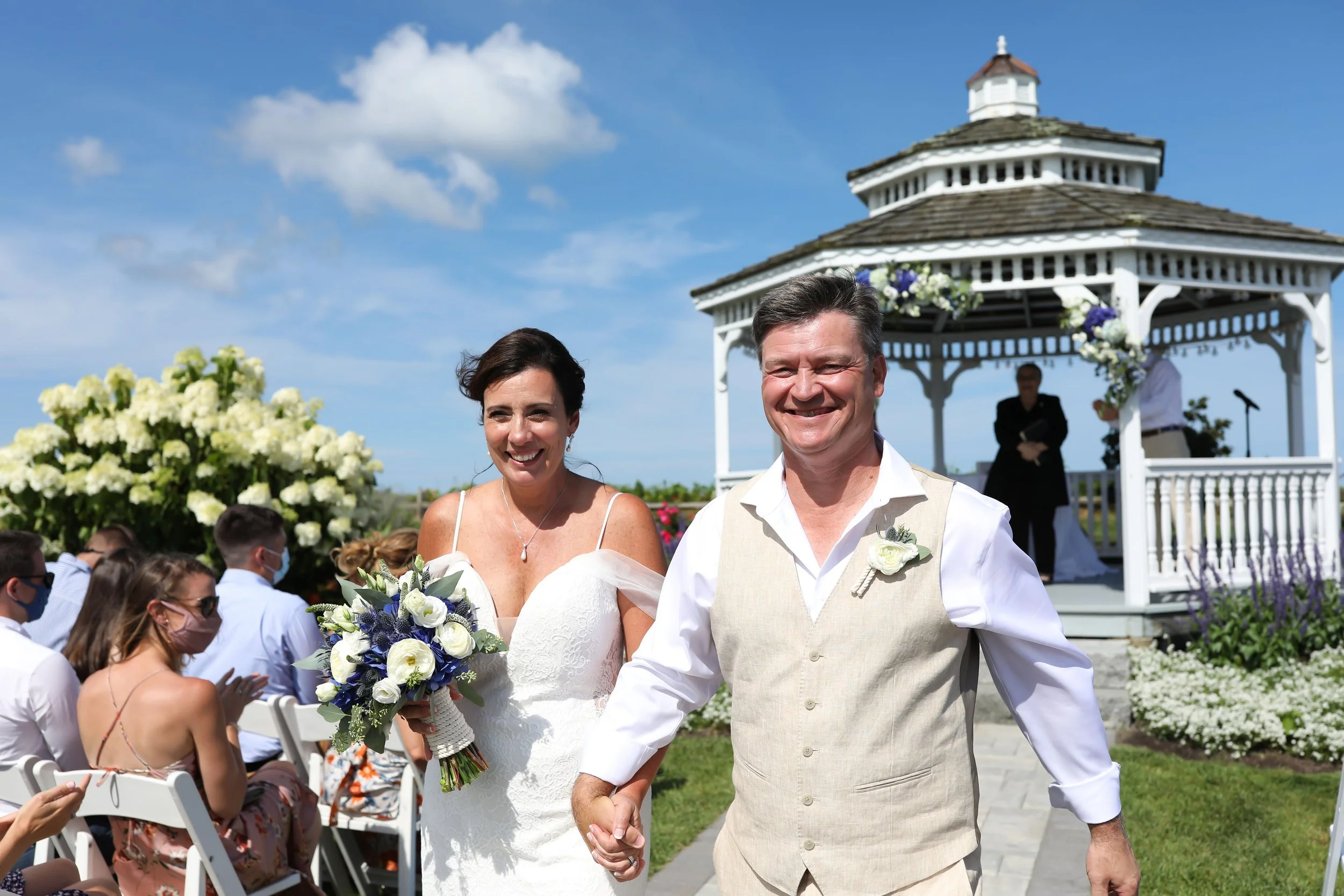A bride and groom holding hands and smiling at an outdoor wedding ceremony on a sunny day, with guests seated and a white gazebo decorated with flowers in the background.
