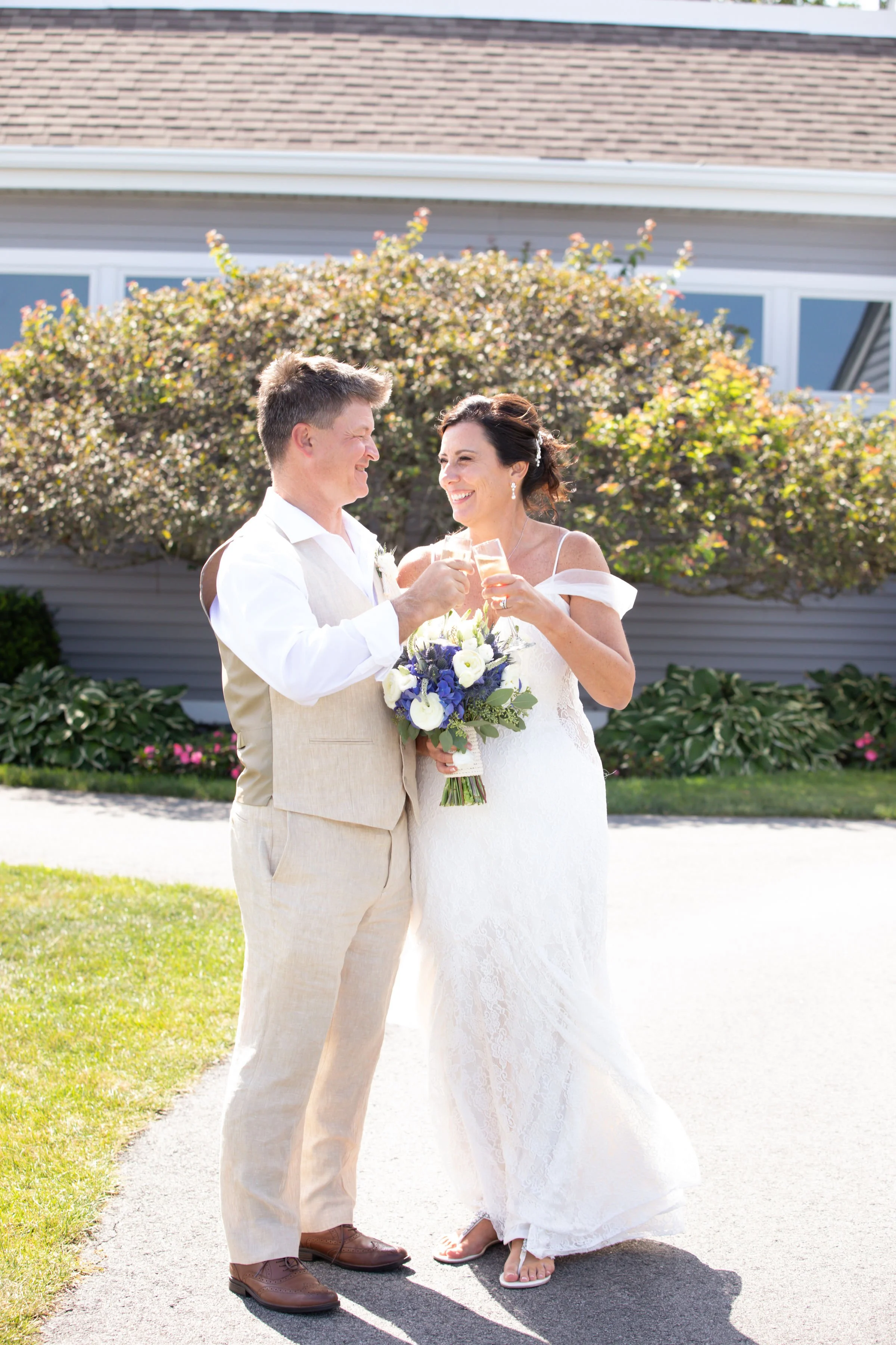 A bride and groom in wedding attire celebrating outdoors, clinking glasses, with the bride holding a bouquet of white and purple flowers, in front of a house with shrubbery and a large tree.