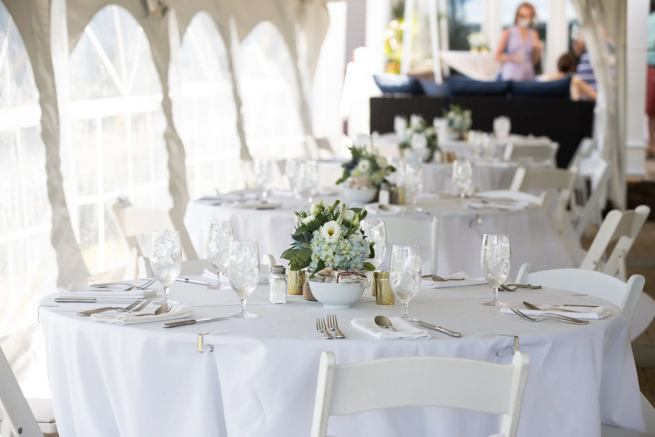Elegant round dining table set with white tablecloth, floral centerpiece, multiple wine glasses, plates, forks, knives, and spoons, in a bright tented venue with windows and people in the background.