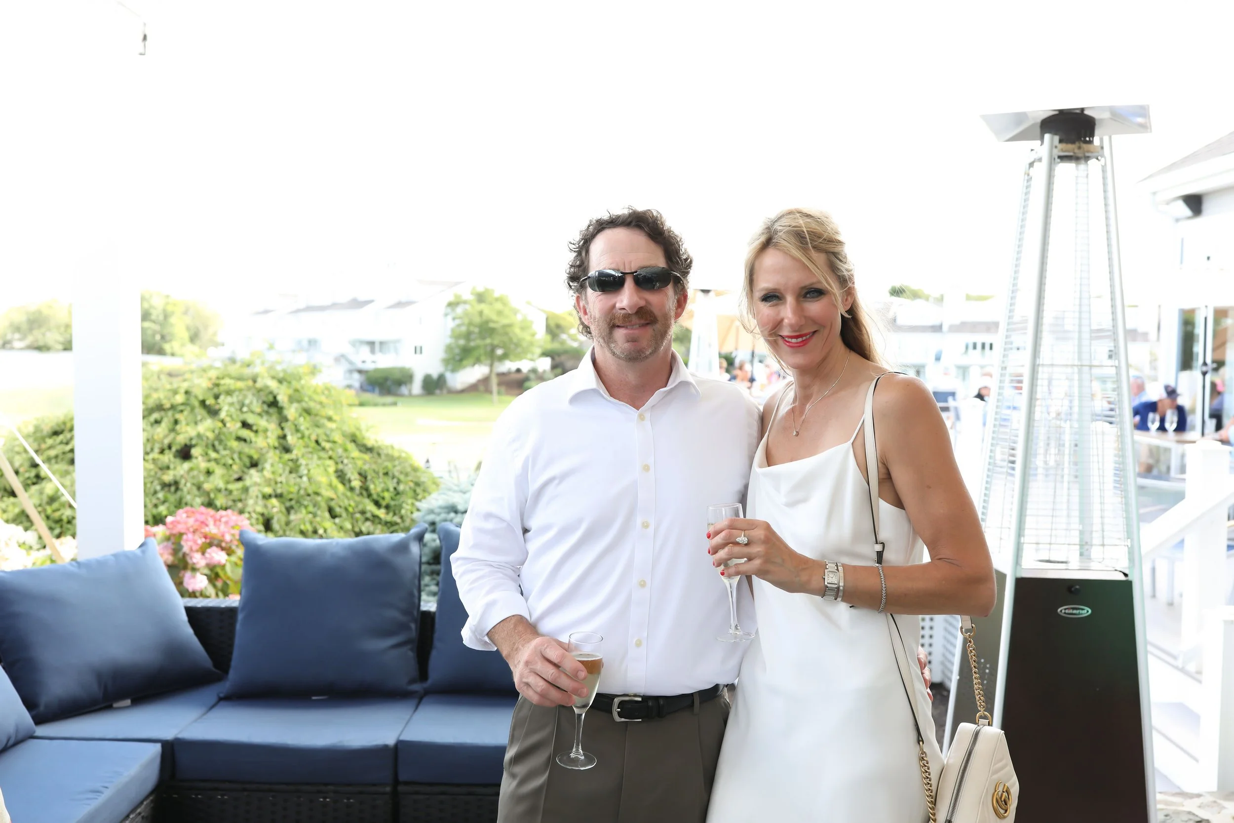 A man and woman standing outdoors, smiling, holding drinks, dressed in formal attire, with outdoor furniture and a patio heater in the background.