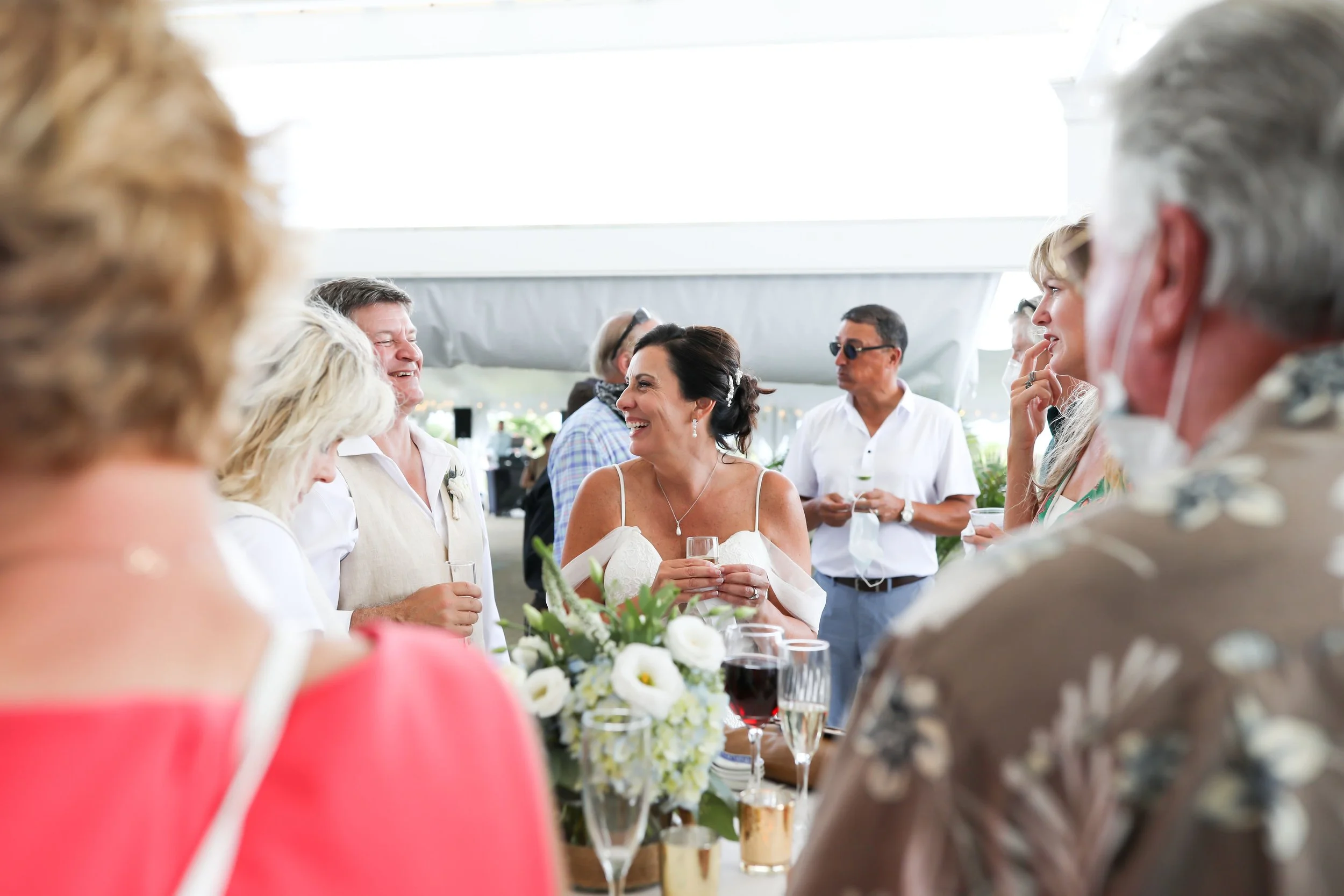 People at a wedding reception, smiling and holding drinks, with a woman in a white dress at the center, surrounded by friends or family in semi-formal attire under a white canopy.