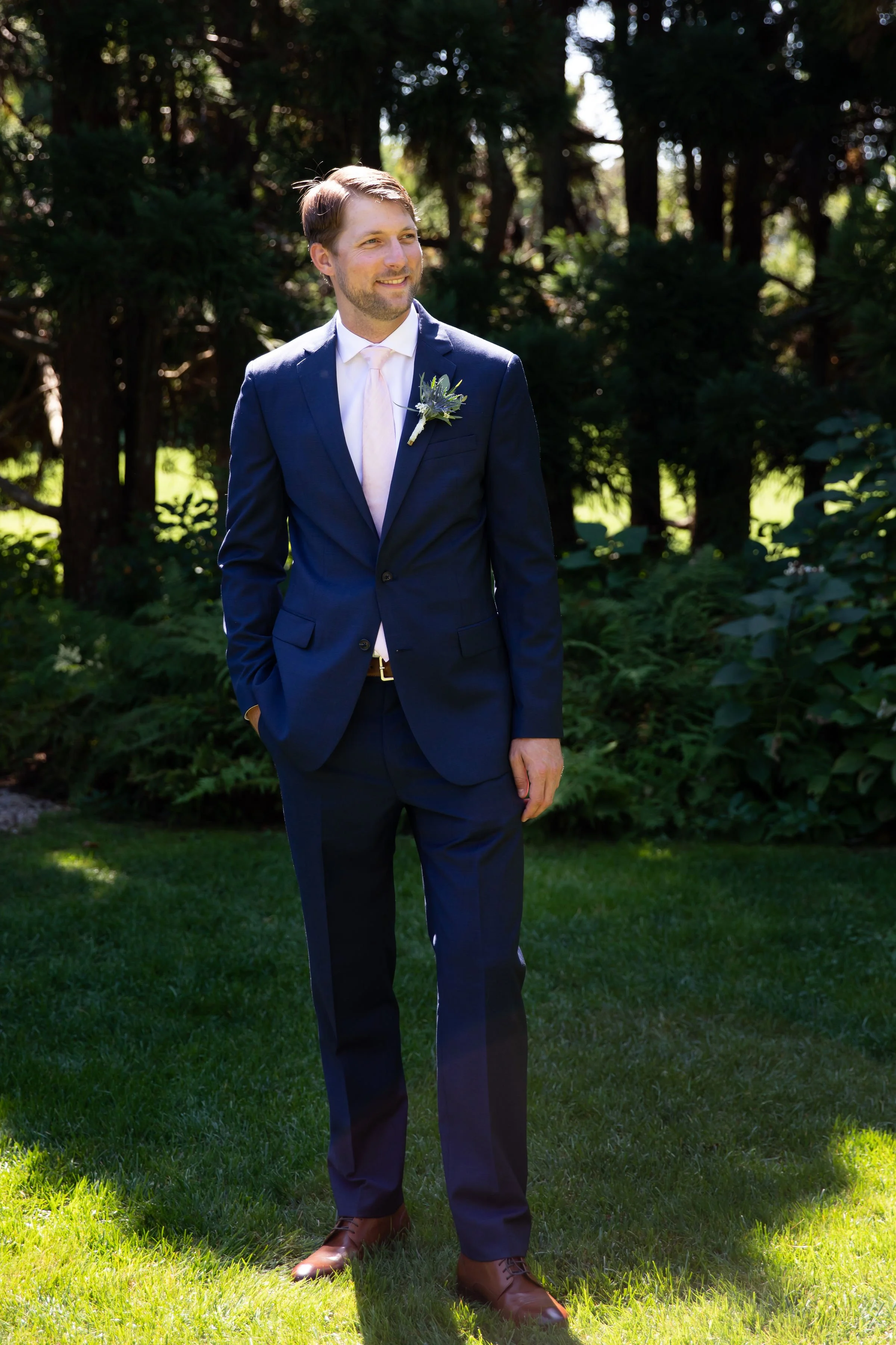 A man in a navy blue suit with a white shirt, light pink tie, and brown shoes stands on grass in a wooded outdoor setting, smiling and looking to the side.