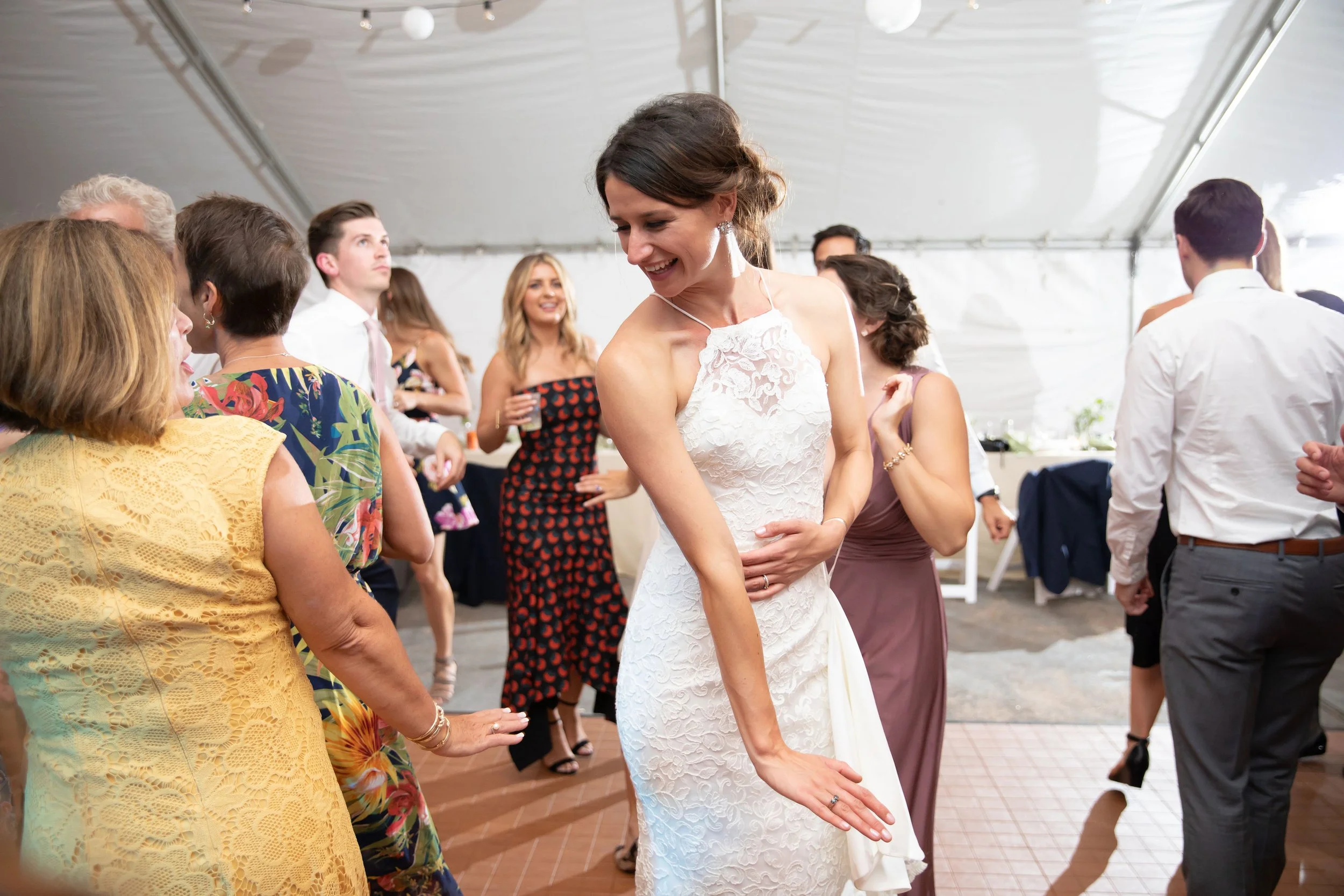 A woman in a white lace wedding dress dancing at a wedding reception surrounded by other guests in colorful dresses and formal attire.