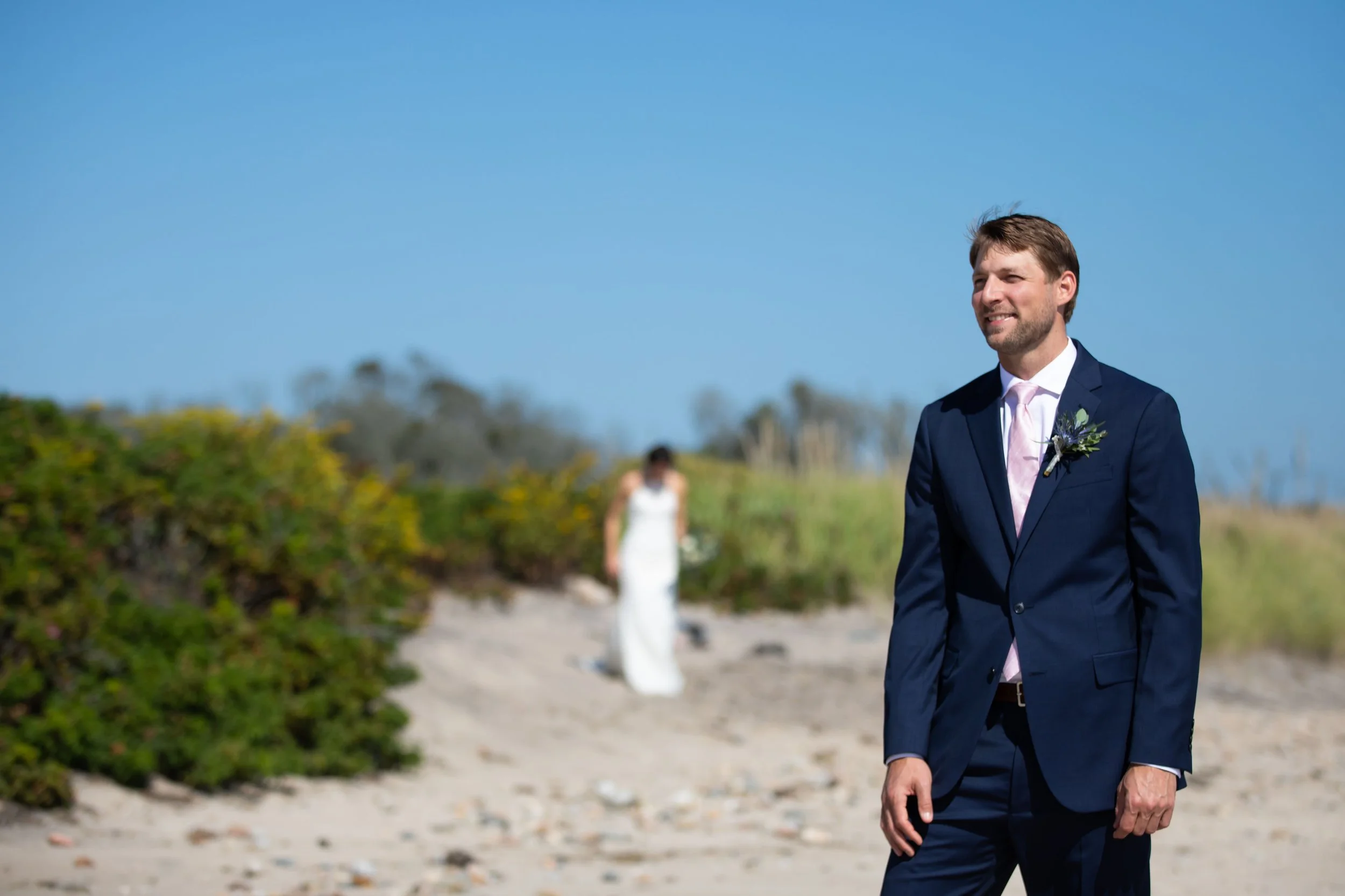 A groom in a navy suit and pink tie standing on a beach, smiling, with a bride in a white wedding dress in the background