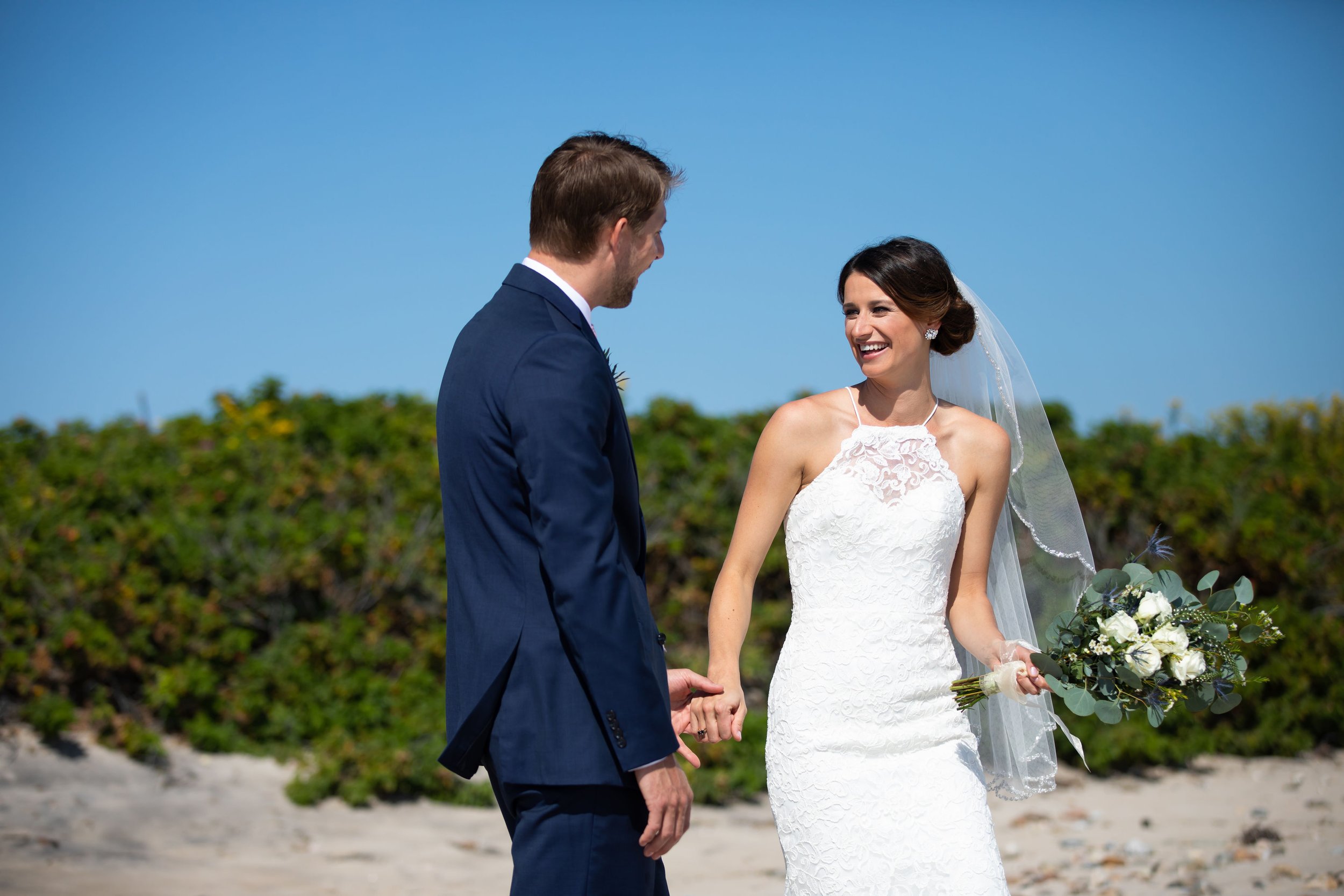 A bride and groom holding hands on a beach, smiling at each other with a background of green bushes and a blue sky.