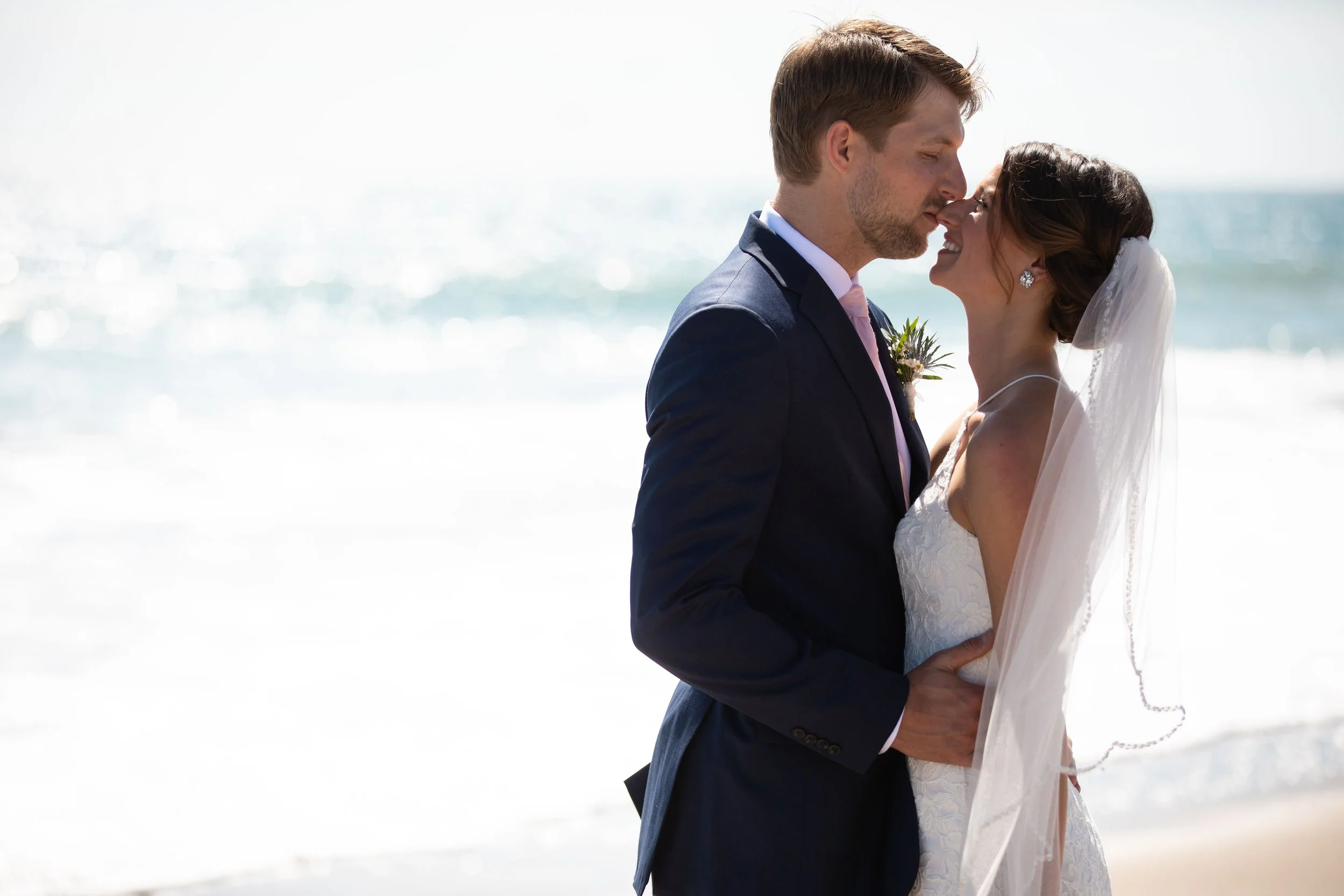 A bride and groom standing close on a beach, foreheads touching, with the ocean in the background.