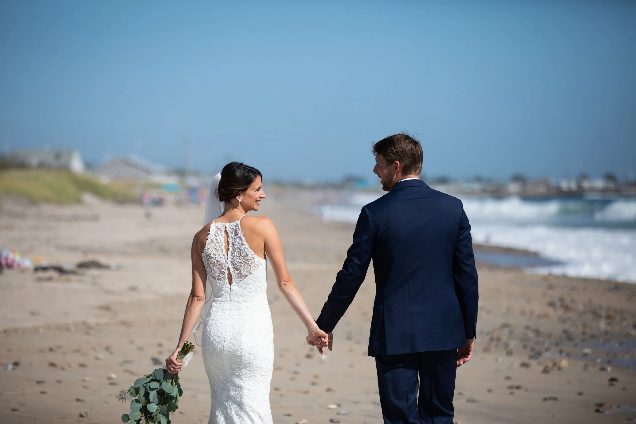 Bride and groom holding hands on the beach, smiling, with the ocean and sky in the background