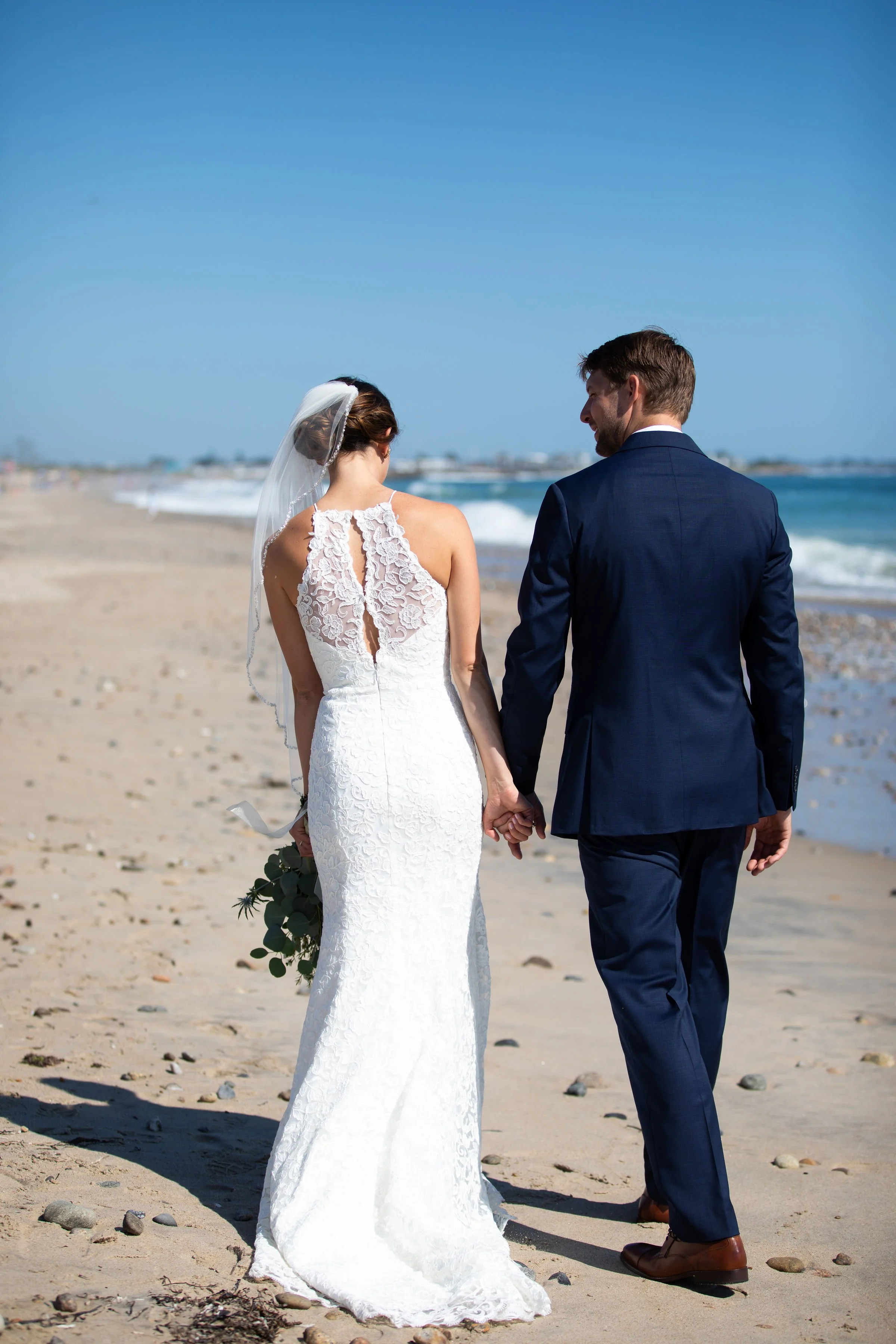 A bride and groom walking hand in hand on the beach during their wedding, with the ocean and blue sky in the background.