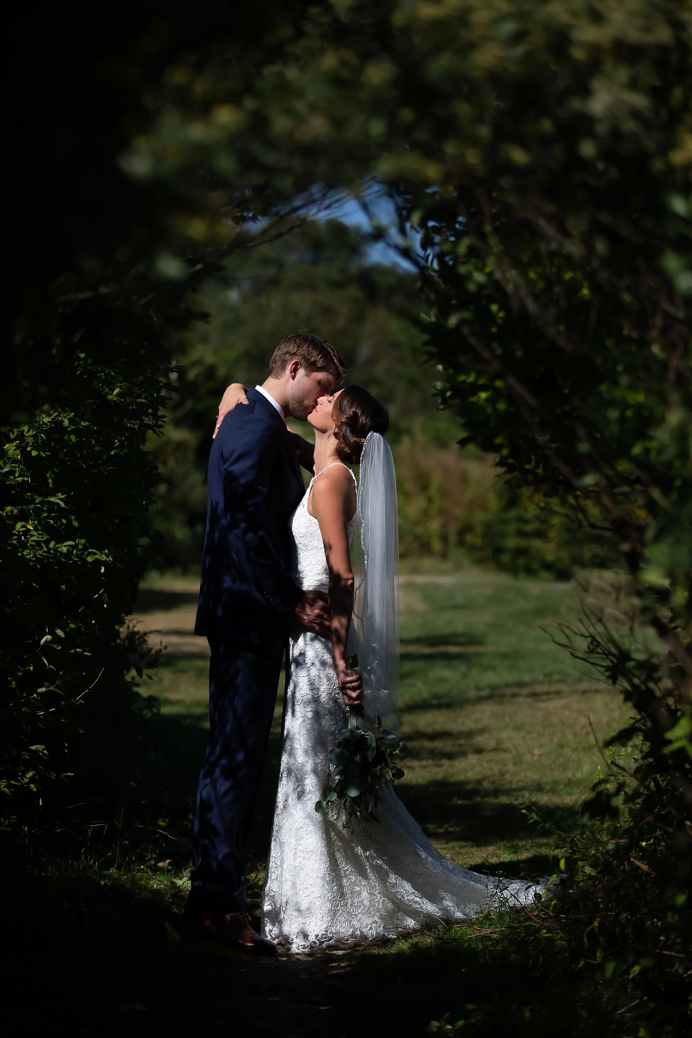 A newlywed couple sharing a romantic kiss outdoors, framed by greenery with shadows and dappled light