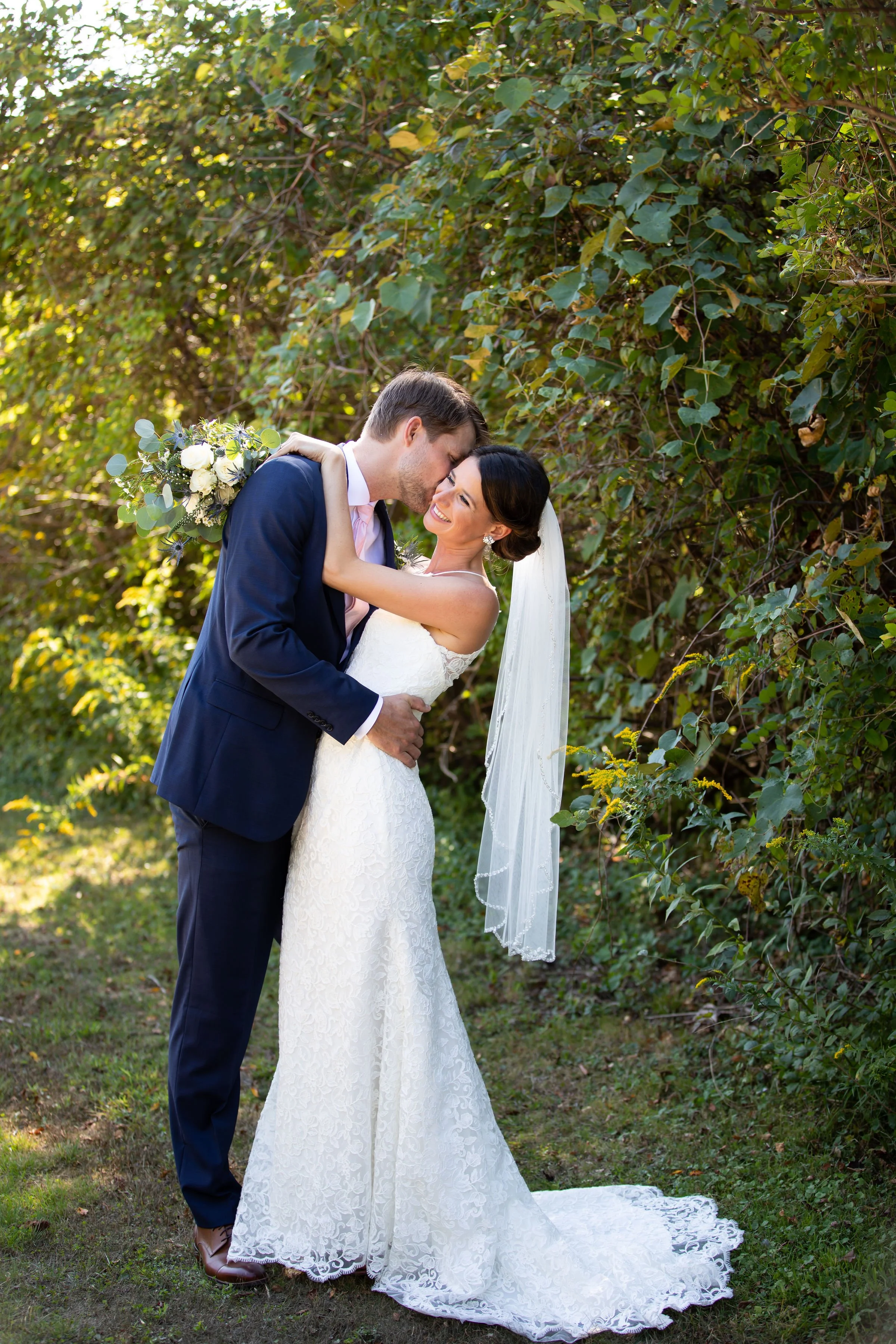 A newlywed couple sharing a romantic moment outdoors, with the groom gently holding the bride's waist and the bride smiling while embracing the groom. They are standing on grass with green leafy bushes in the background, dressed in wedding attire.
