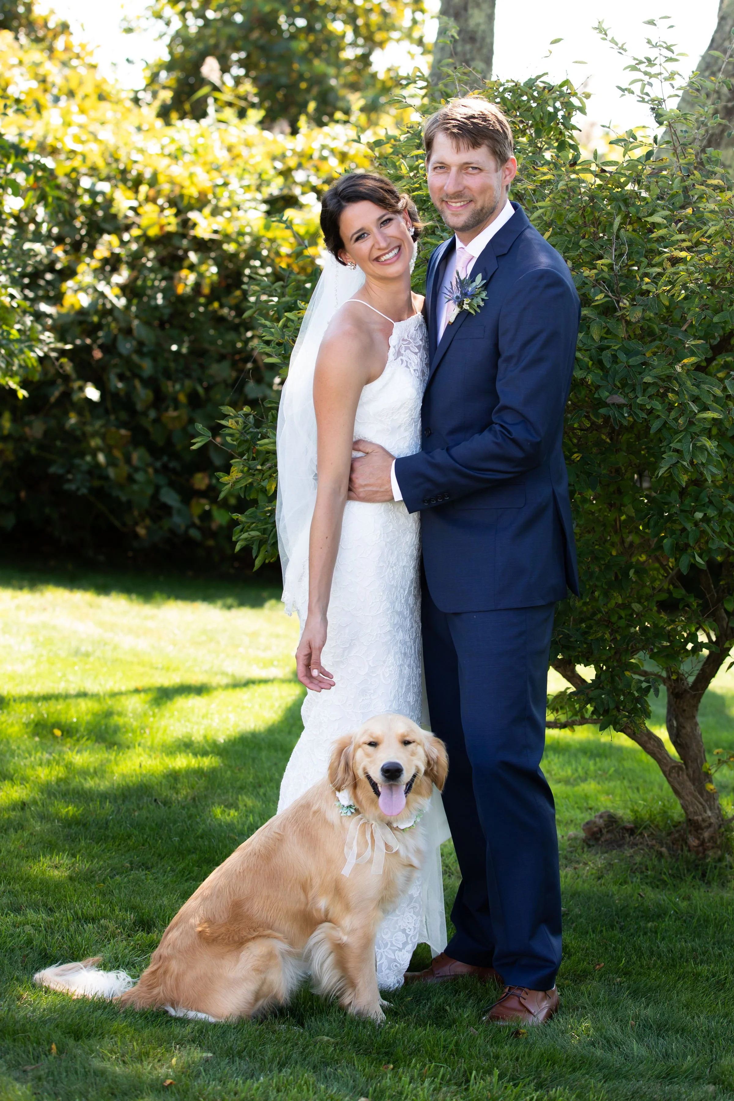 A newlywed couple smiling and posing outdoors on a sunny day, with a golden retriever dog sitting in front of them on green grass and lush bushes in the background.