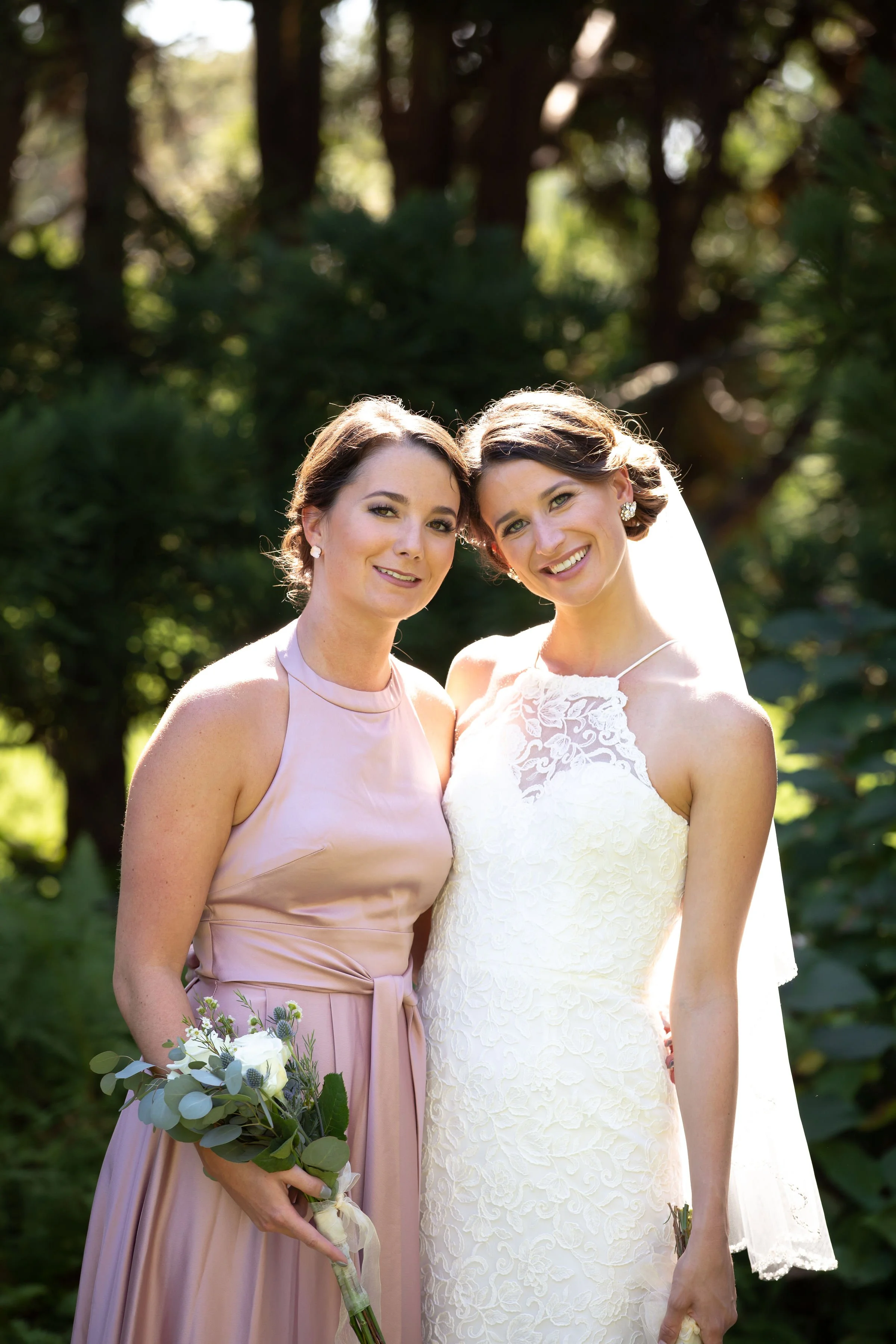 A bride and woman standing together outdoors, smiling, in a wooded area. The bride is wearing a white lace wedding dress and veil. The other woman is in a pink dress and holding a bouquet.
