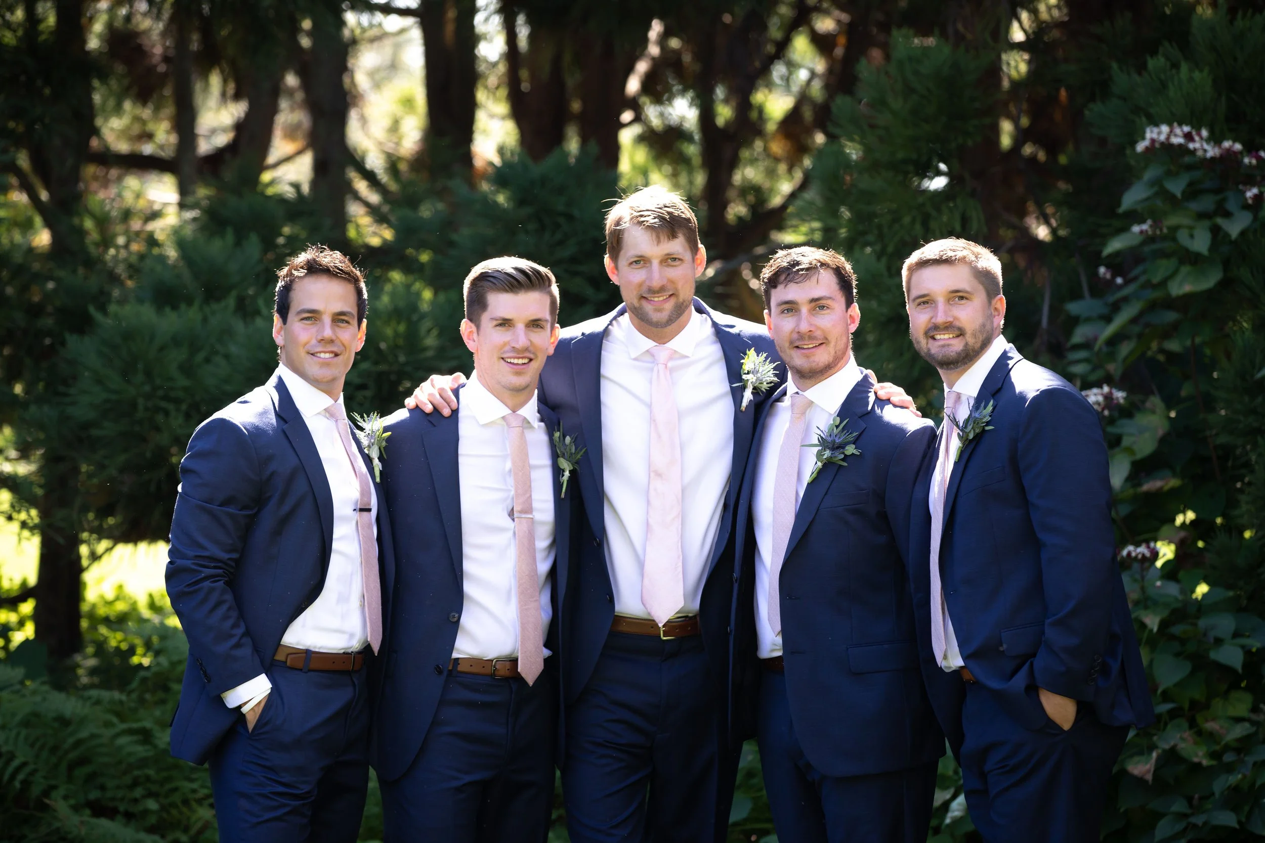 Six men dressed in dark suits, white shirts, and light pink ties, standing outdoors with trees in the background. They are smiling, with some placing their hands on each other's shoulders, celebrating at a wedding.