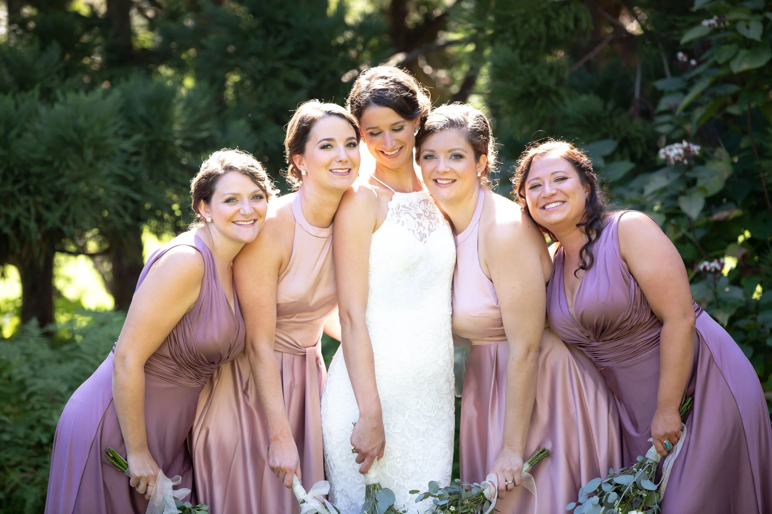 A bride and four bridesmaids smiling outdoors, with the bridesmaids wearing lavender dresses and holding bouquets, and the bride in a white lace gown.