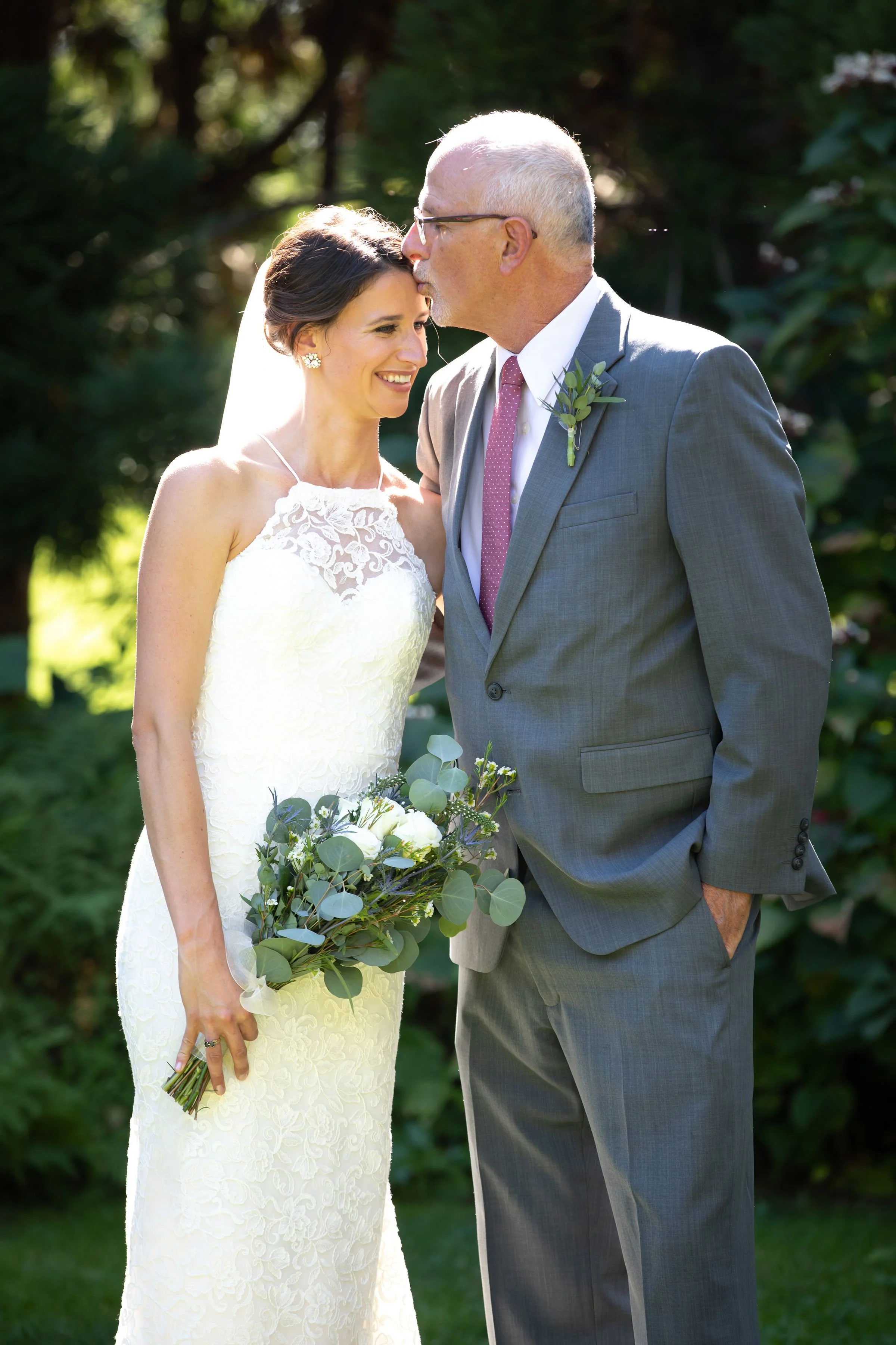 A bride and an older man, likely her father, sharing a moment outdoors. The bride is in a white lace wedding gown, holding a bouquet of white flowers and eucalyptus leaves. The man is dressed in a gray suit with a pink tie, kissing the bride on her f