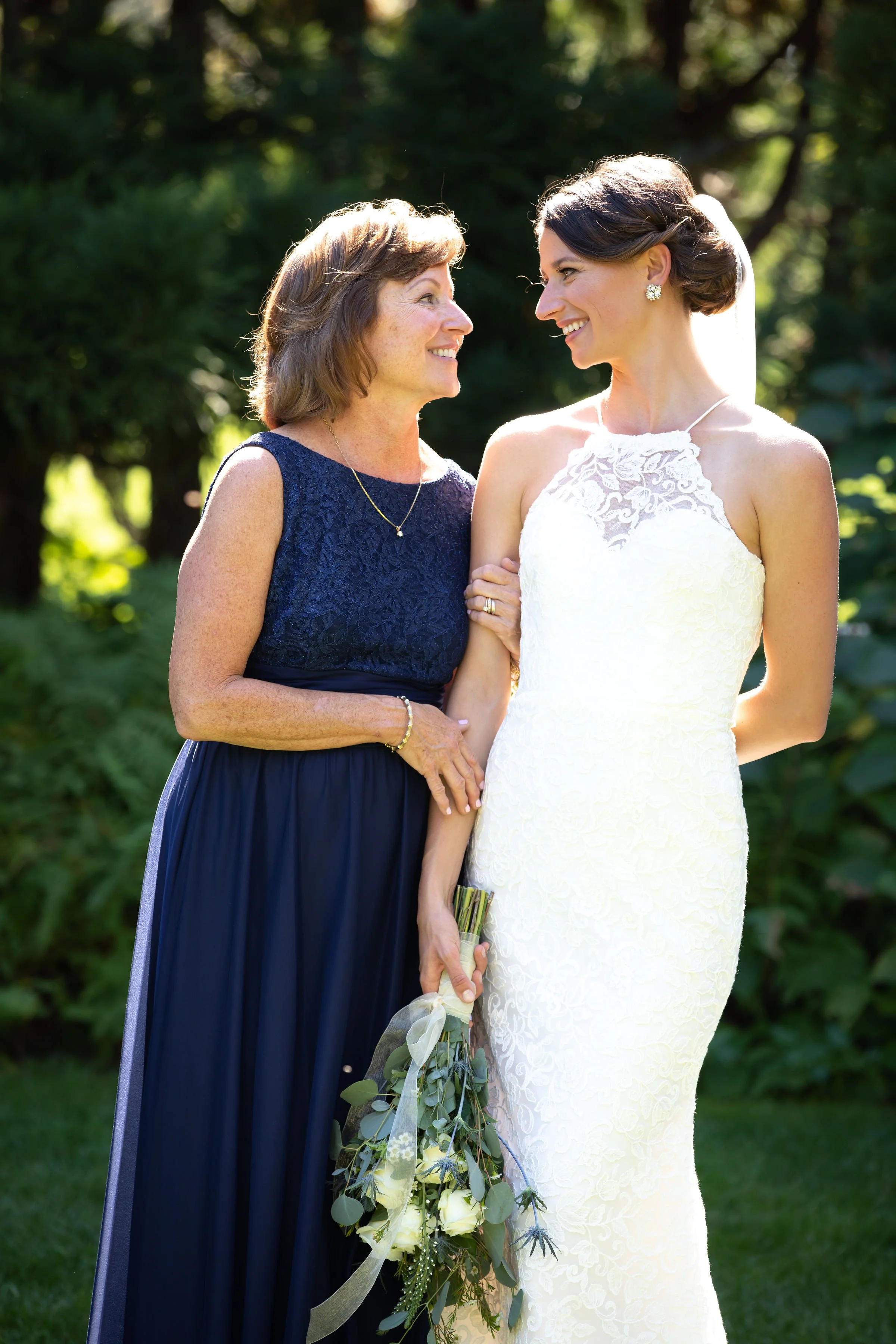 A bride in a white lace wedding dress holding a bouquet, smiling at an older woman in a navy blue dress, outdoors with greenery in the background.