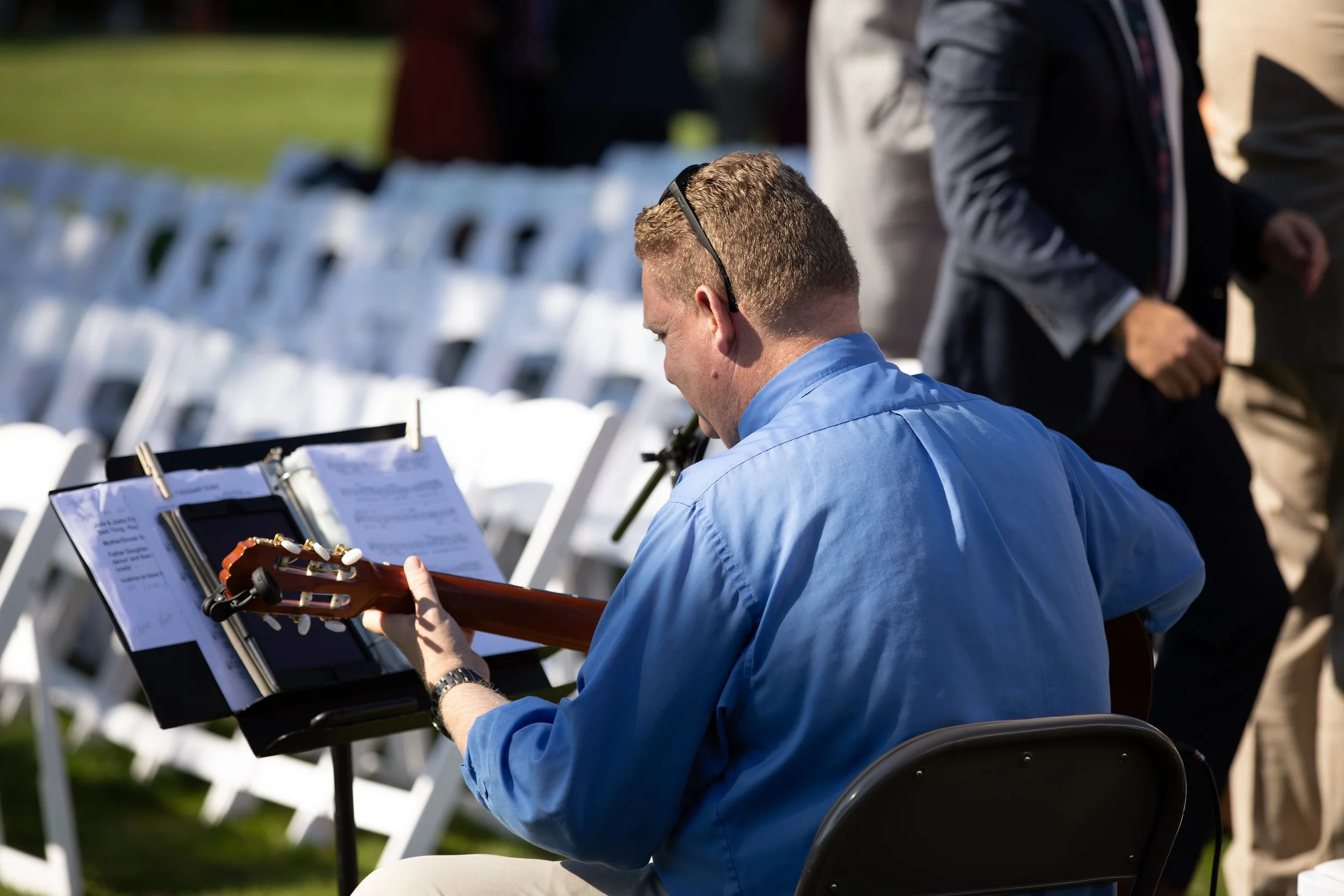 A man in a blue shirt playing guitar at an outdoor event with rows of empty white chairs behind him.