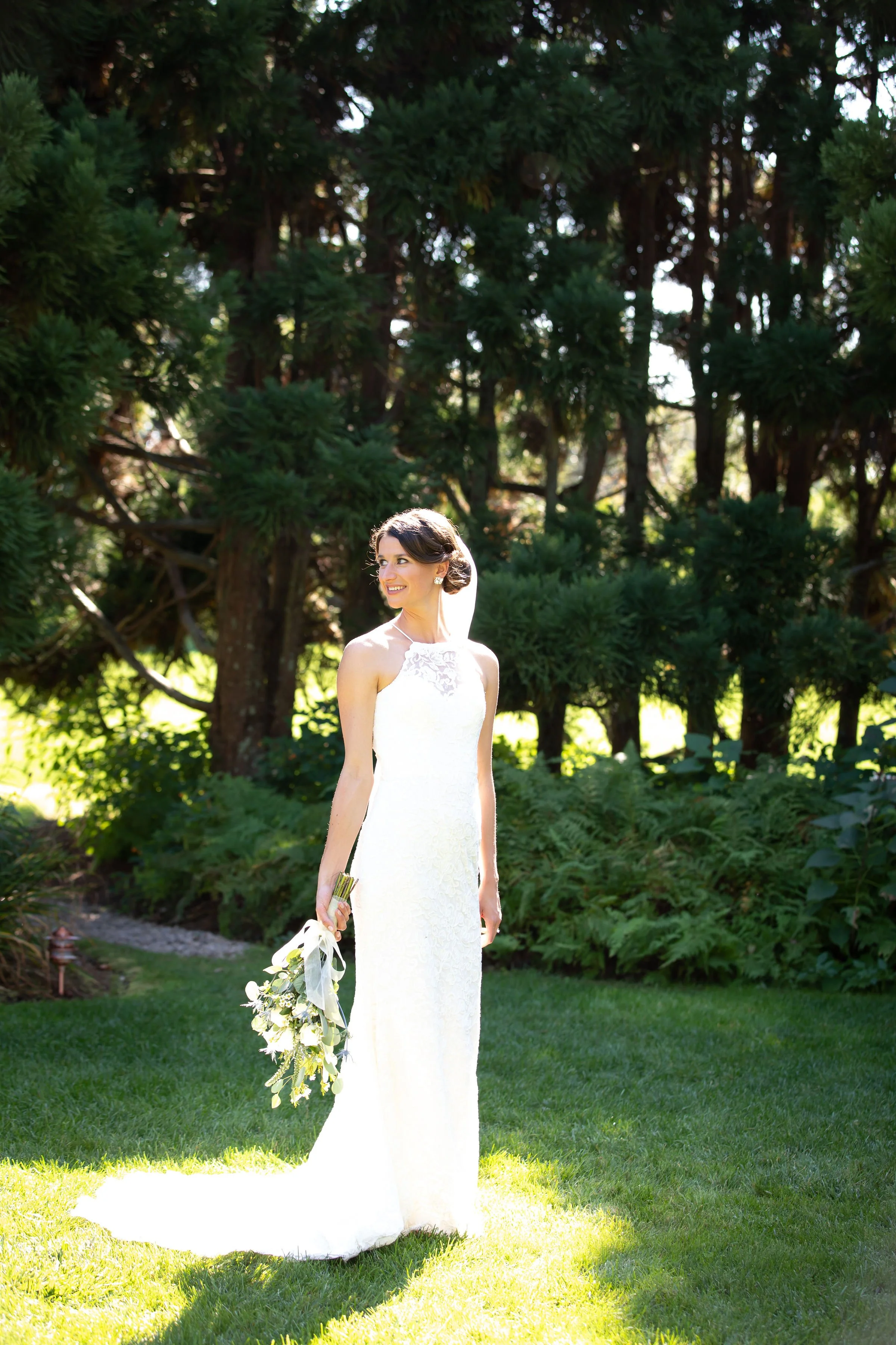 A bride in a white wedding dress holding a bouquet stands on a grassy lawn with lush green trees in the background, smiling and looking to the side.