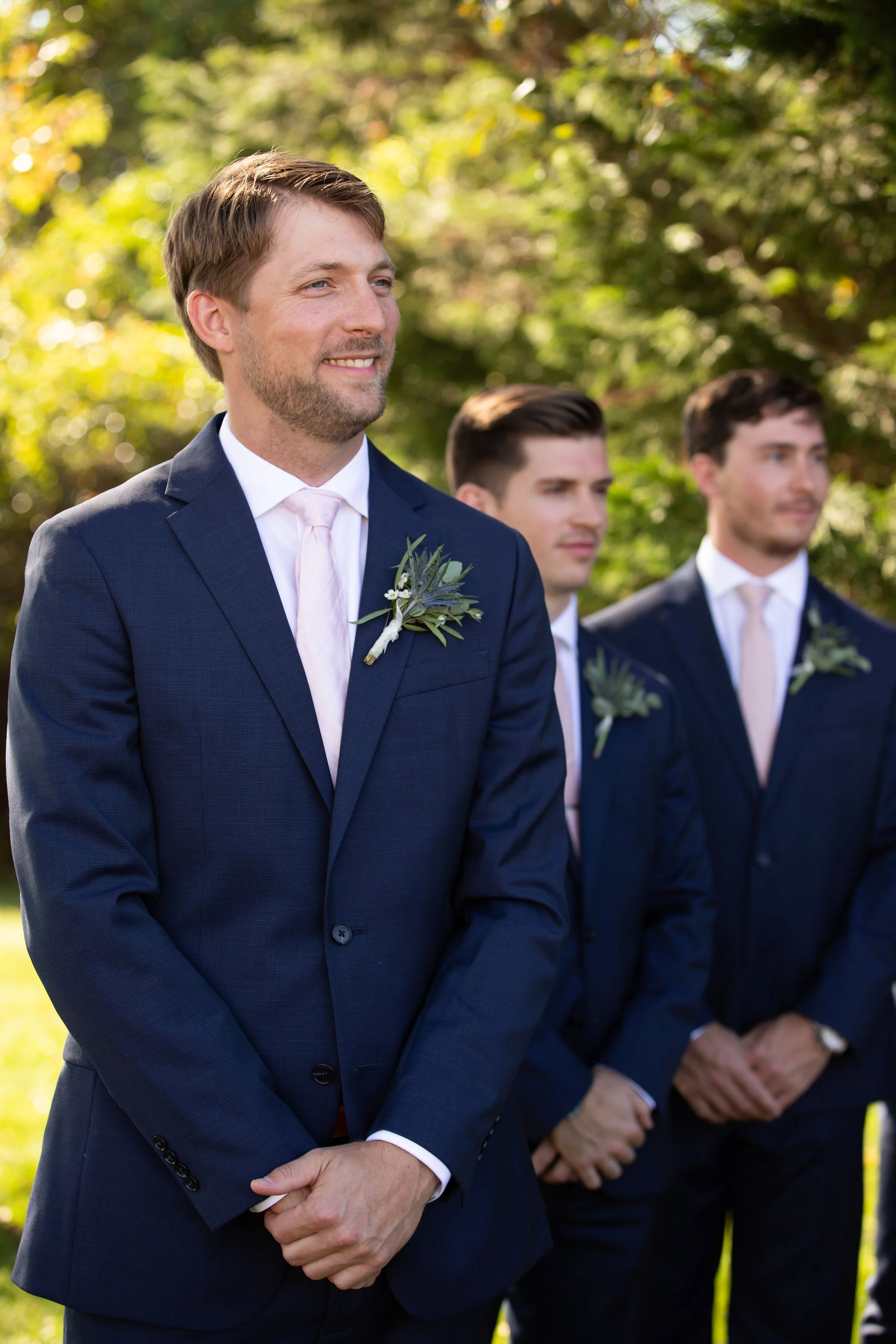 A man in a dark blue suit with a pink tie and boutonniere standing outdoors at a wedding, smiling, with two other men in similar attire behind him.