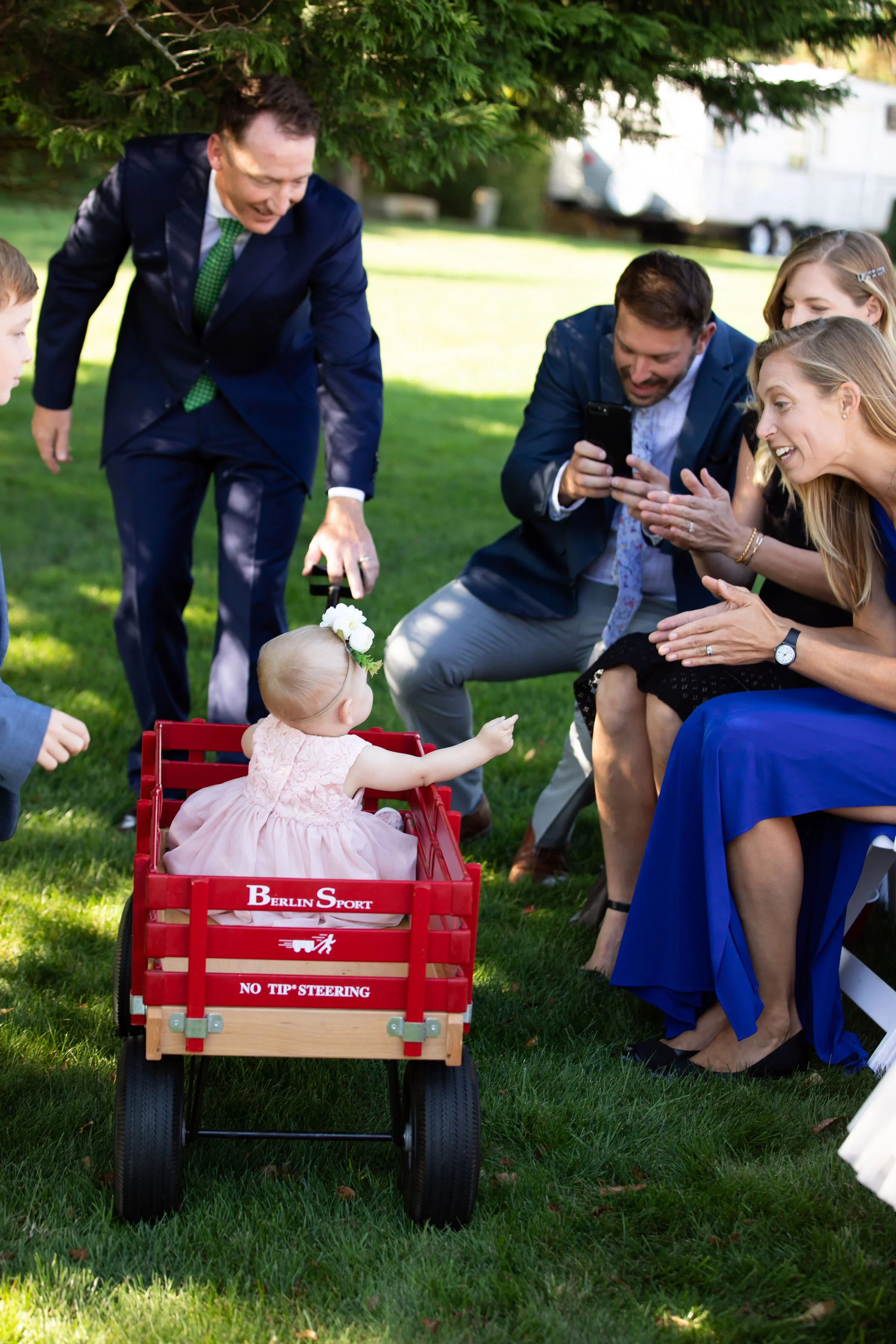 A group of adults in formal attire gathered outdoors, excitedly taking photos of a young girl in a pink dress sitting in a red wagon with 'Berlin Sport' and 'No Tip Steering' written on it, under a green tree.