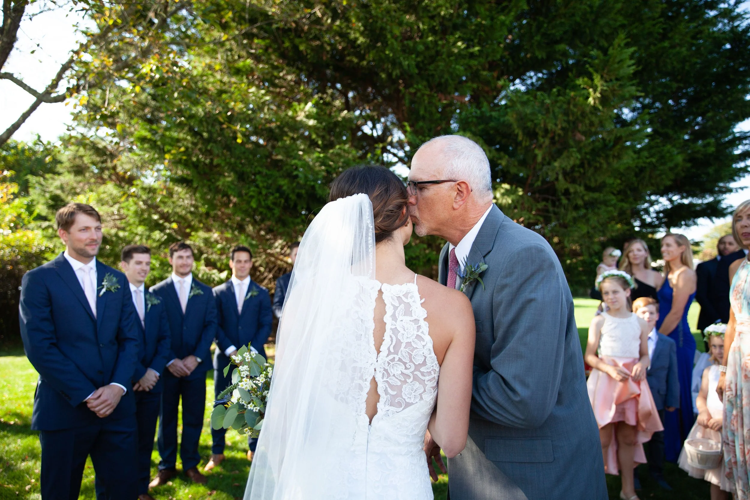A bride and an older man, likely her father, sharing a kiss during an outdoor wedding ceremony. The bride is wearing a white lace wedding dress and a veil, holding a bouquet. Guests in formal attire are seen in the background, with some children, all