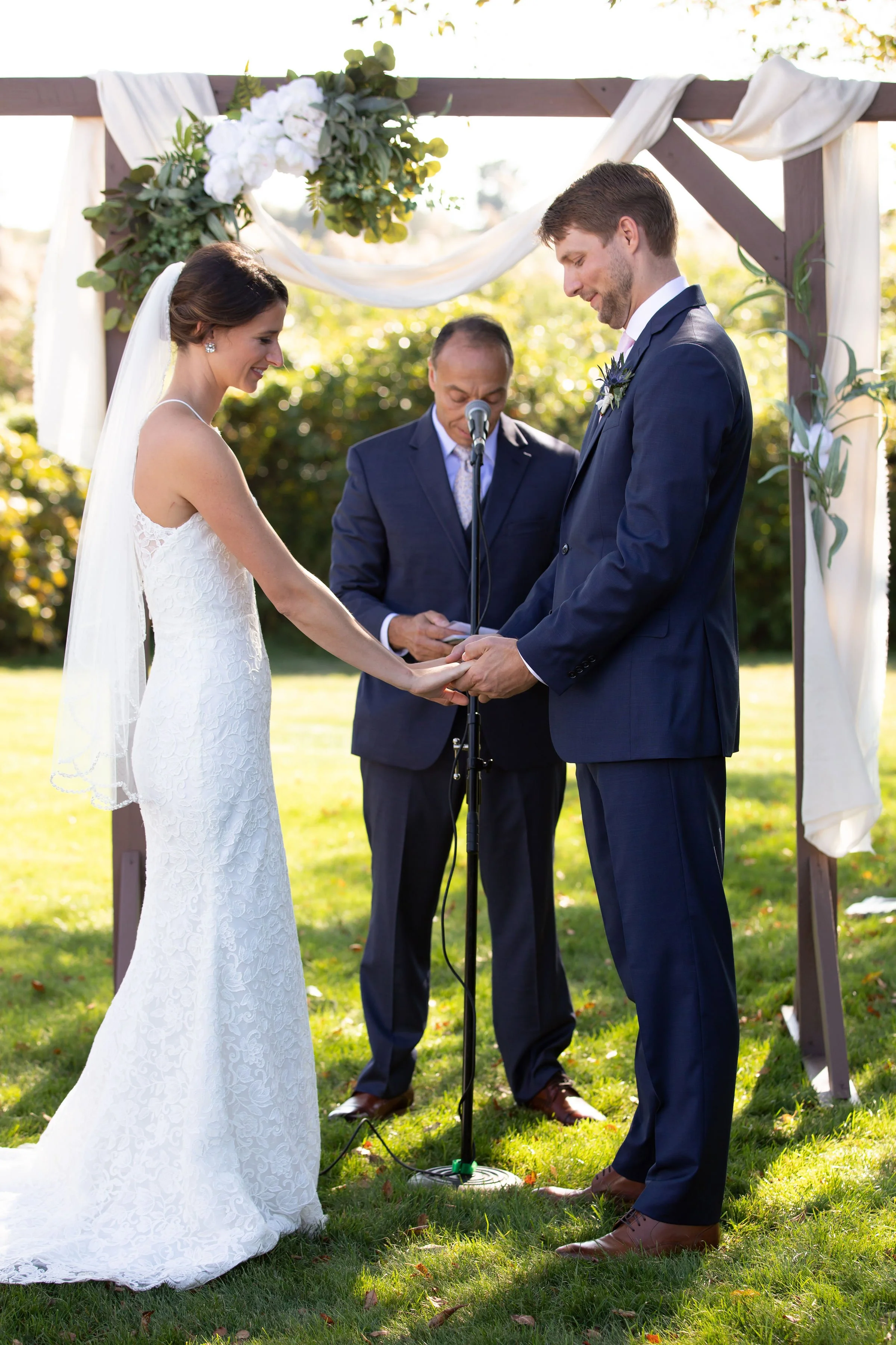 A bride and groom holding hands during their outdoor wedding ceremony under a wooden arch decorated with white fabric and flowers, with an officiant in the background.