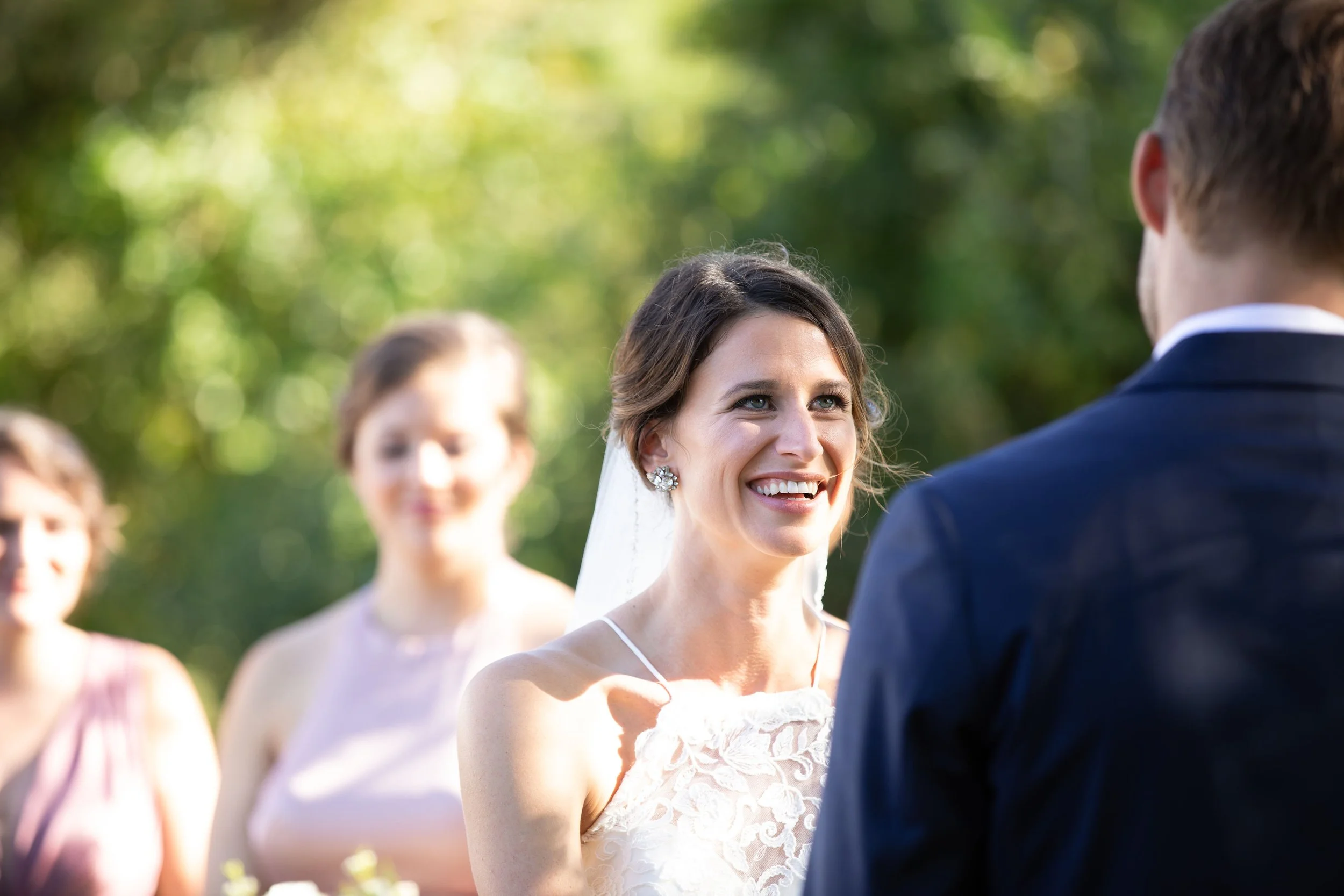 Bride smiling during wedding ceremony outdoors, with groom facing her and bridesmaids in the background.
