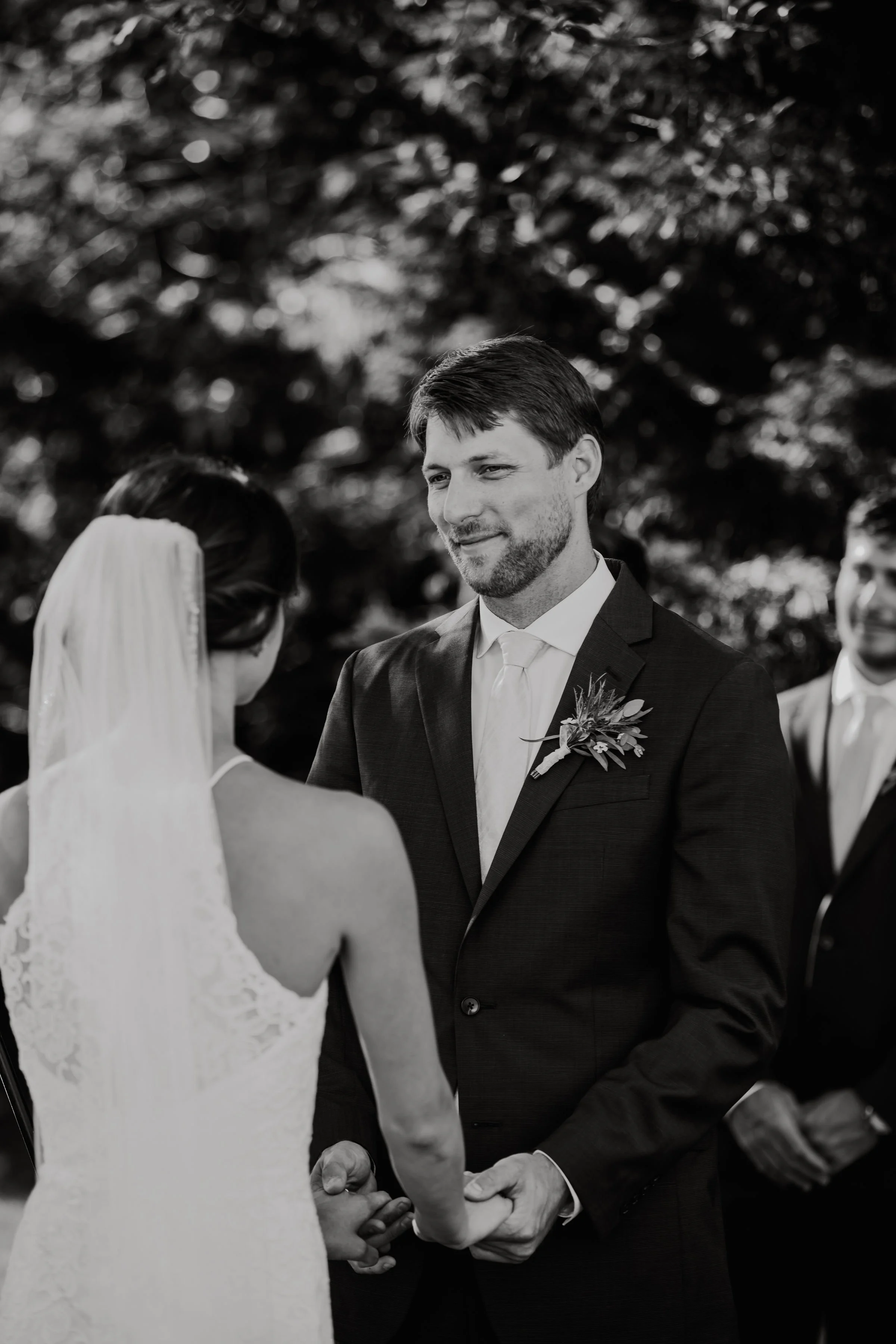Black and white photo of a wedding ceremony with a bride and groom holding hands, exchanging vows, outdoors with trees in the background.