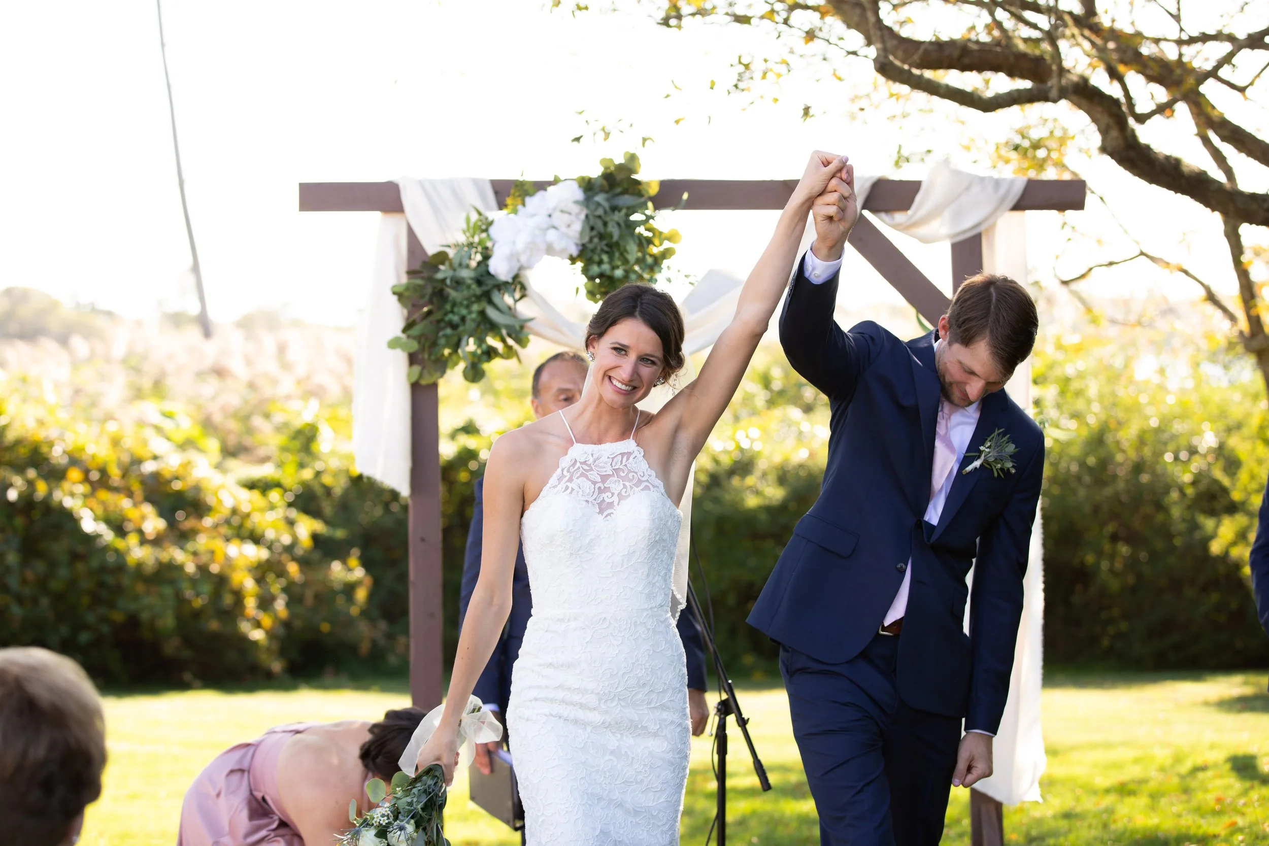 A bride and groom celebrate their wedding outdoors, with the bride smiling and raising her arm as the groom bows his head, set against a garden backdrop with a decorated wooden arch.