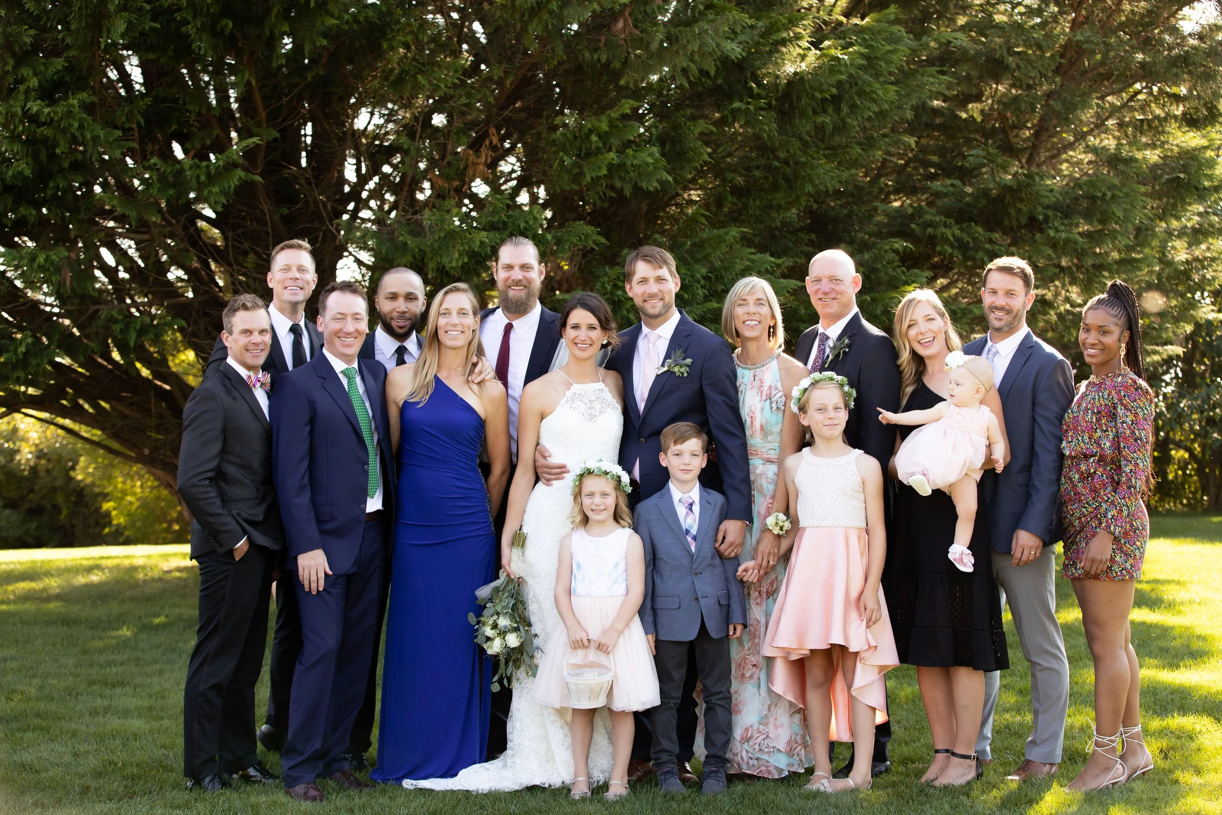 Group of people at a wedding, including the bride and groom, family, and friends, posing outdoors in front of a large tree on a sunny day.