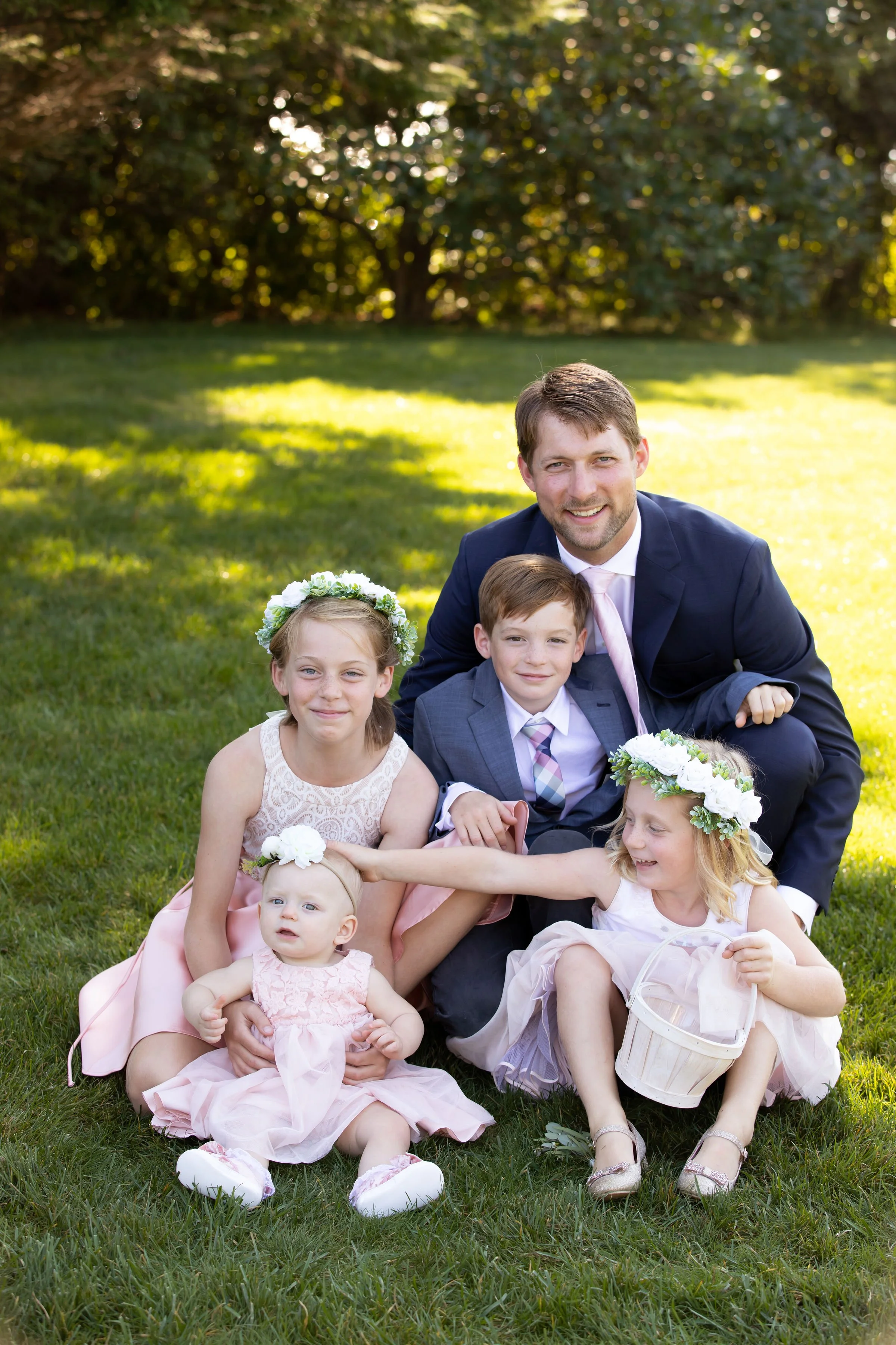 Family of six, including an adult man and five children, sitting on grass in a park, all dressed in formal and semi-formal attire, surrounded by trees and sunlight.
