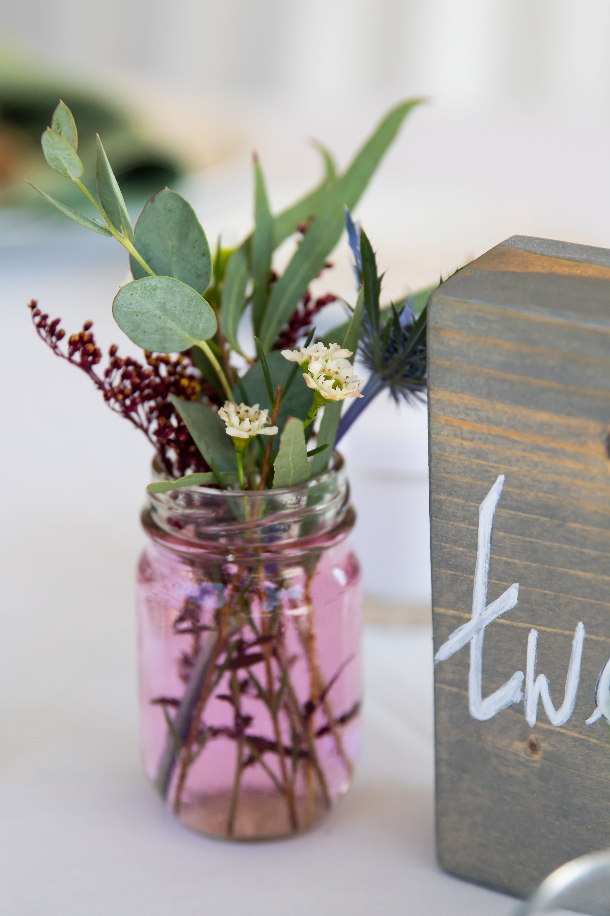 A small bouquet of greenery and white flowers in a light pink glass jar set on a white surface next to a wooden block with white painted letters.