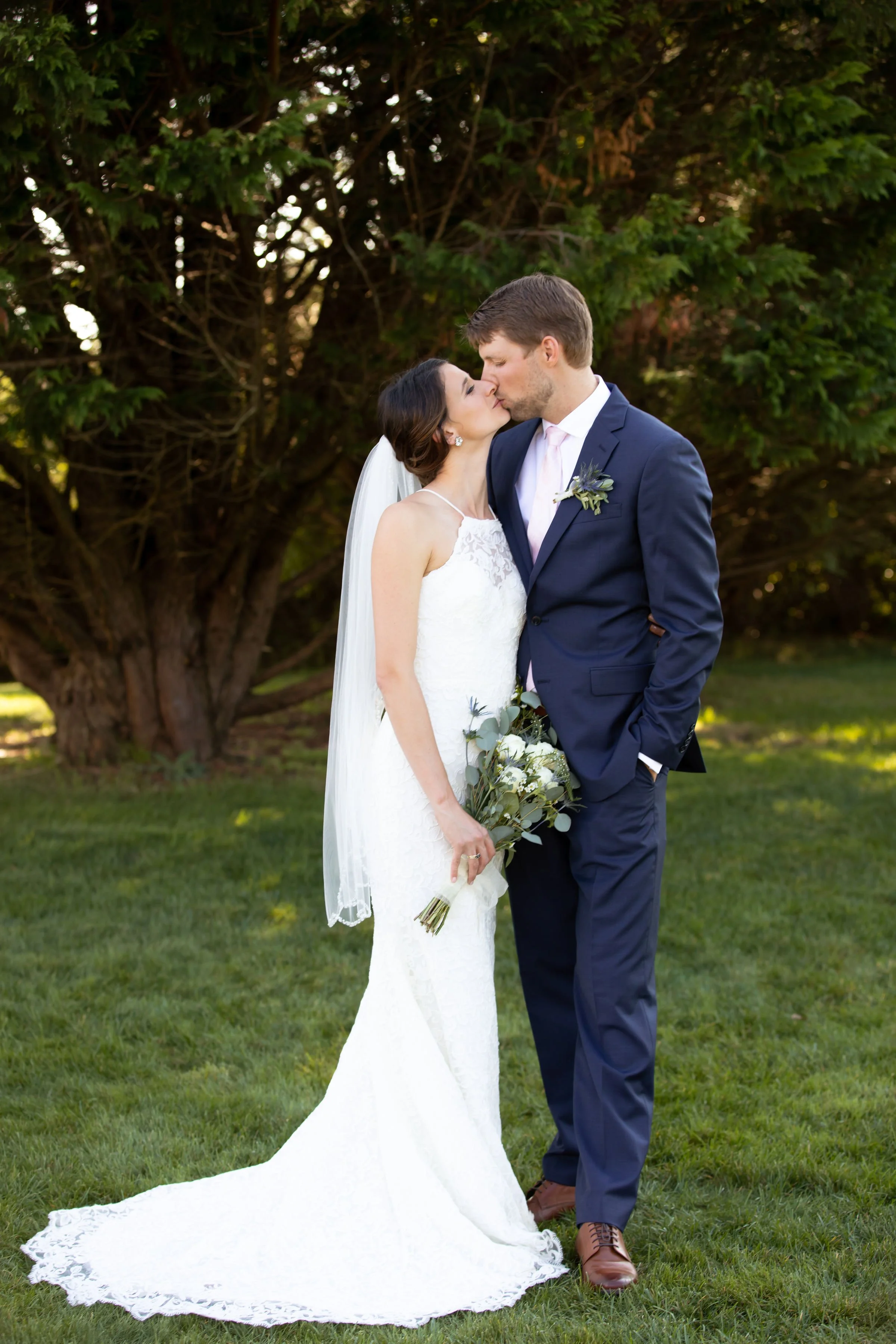 A bride and groom kissing outdoors, the bride in a white lace wedding dress holding a bouquet, and the groom in a navy suit with a boutonniere, standing on grass in front of a large tree.