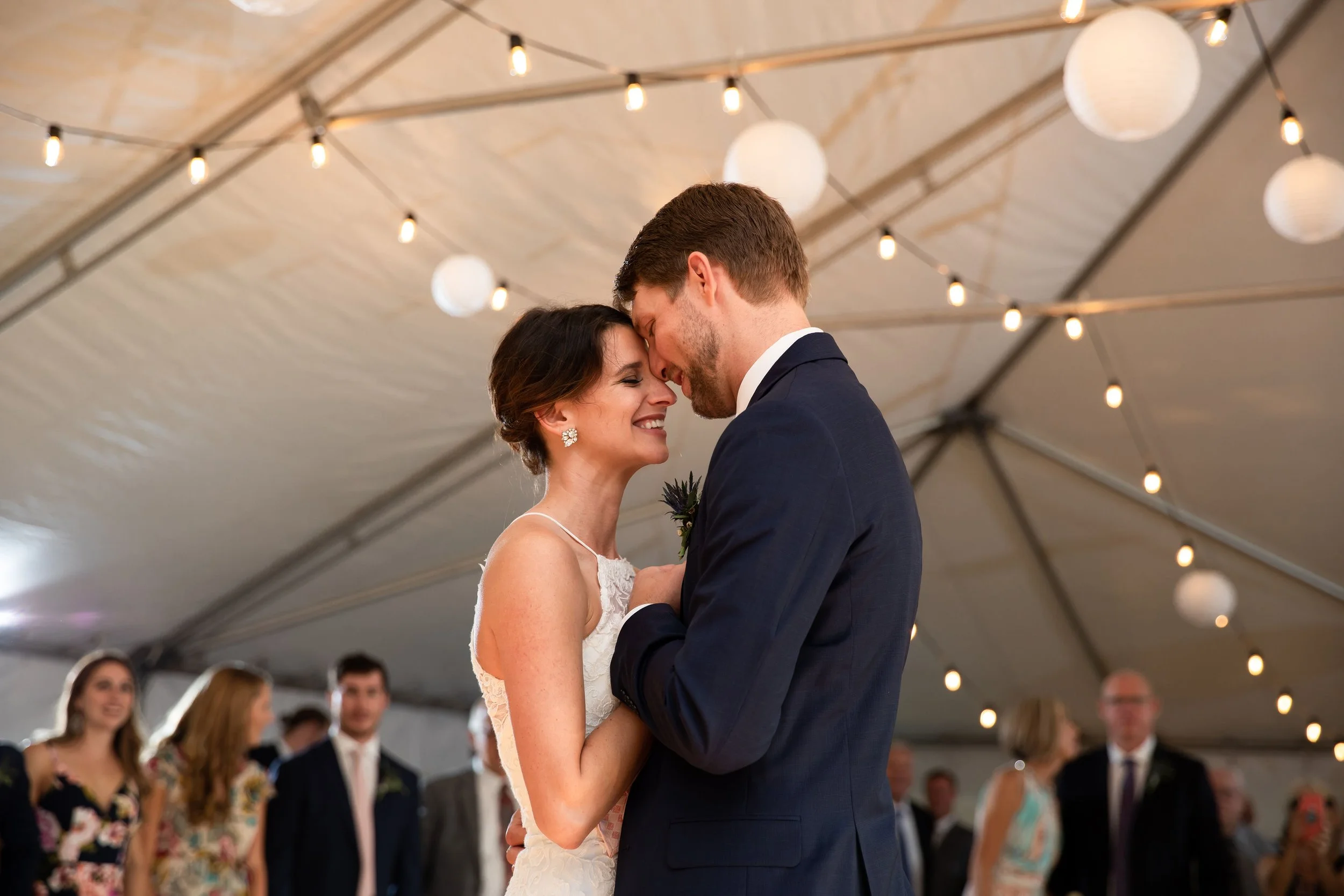 A bride and groom share a dance at their wedding reception, with the bride in a white lace dress and the groom in a navy suit, under hanging string lights and paper lanterns.