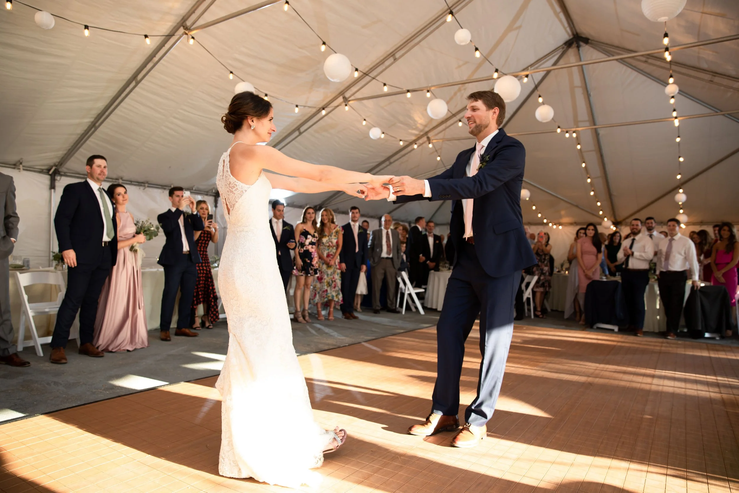 Bride and groom dancing at their wedding reception in a decorated tent with string lights, surrounded by guests.