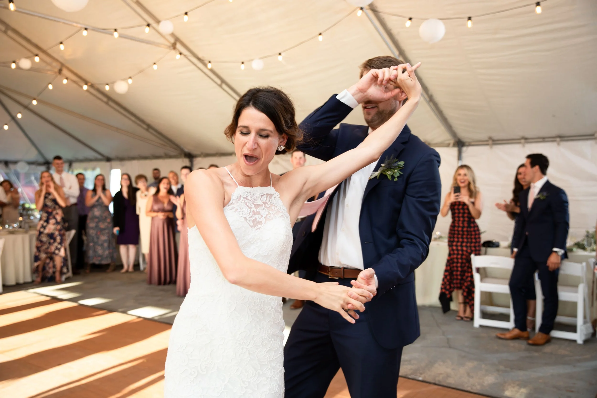 Bride and groom dancing at a wedding reception inside a decorated tent, with guests watching and taking photos in the background.