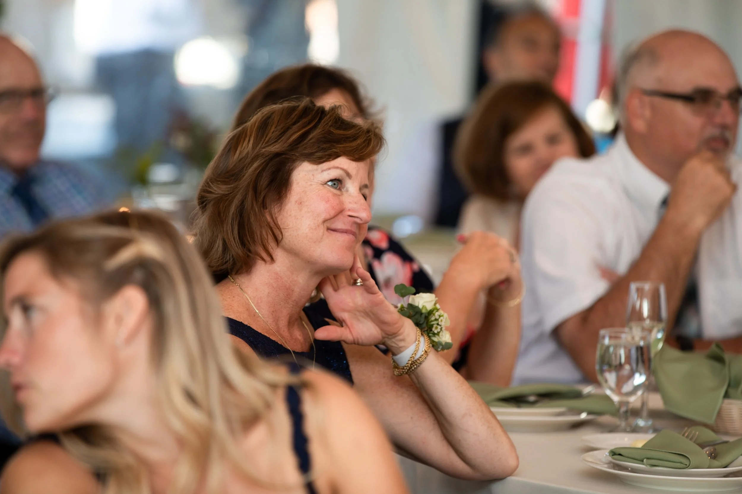 A woman with short brown hair, wearing a navy dress, smiling and listening at a formal gathering or wedding reception, seated at a table with green napkins and glassware.