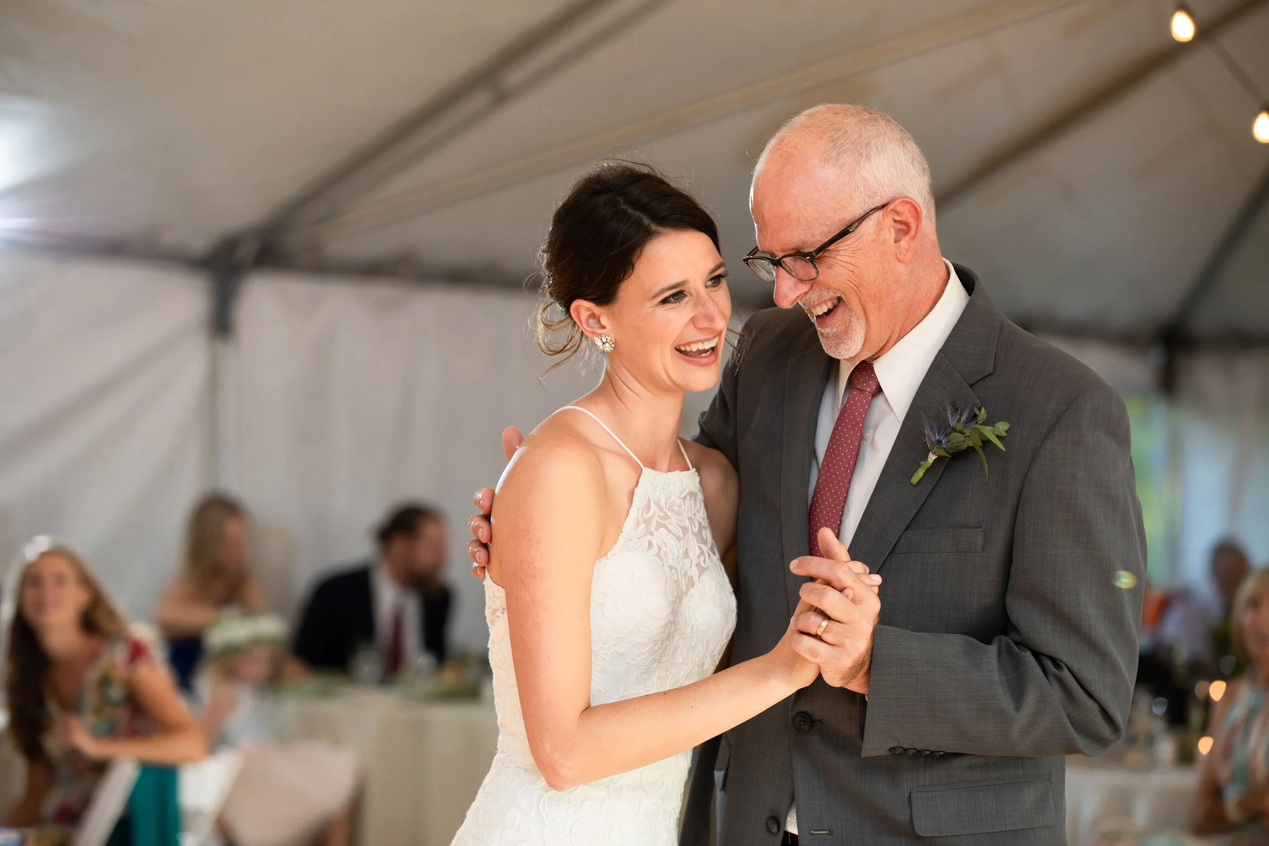 A bride and an older man, possibly her father, sharing a dance at a wedding reception under a large tent, both smiling.