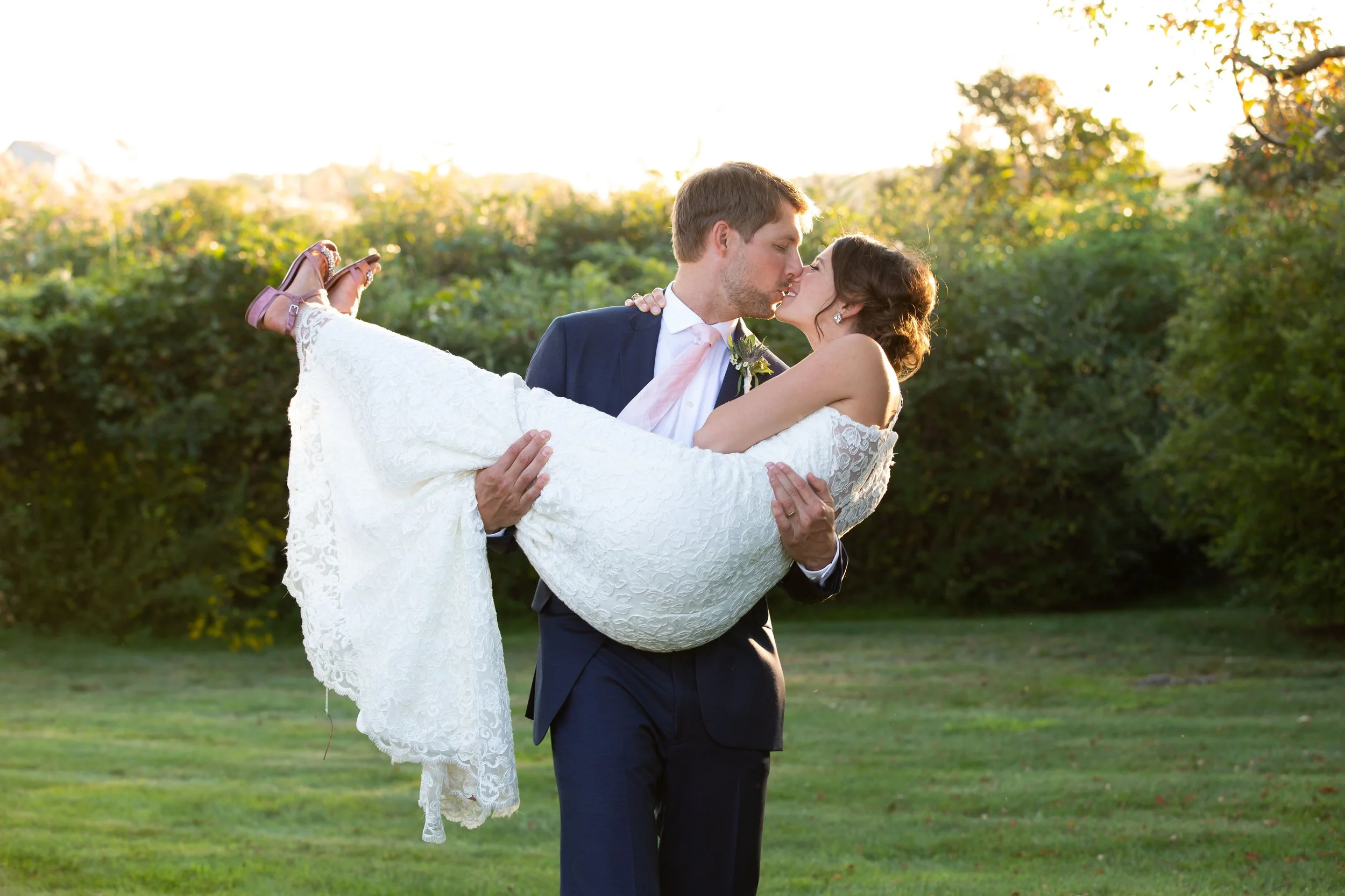 A groom in a dark blue suit holding a bride in a white lace wedding dress outdoors at sunset, both kissing.
