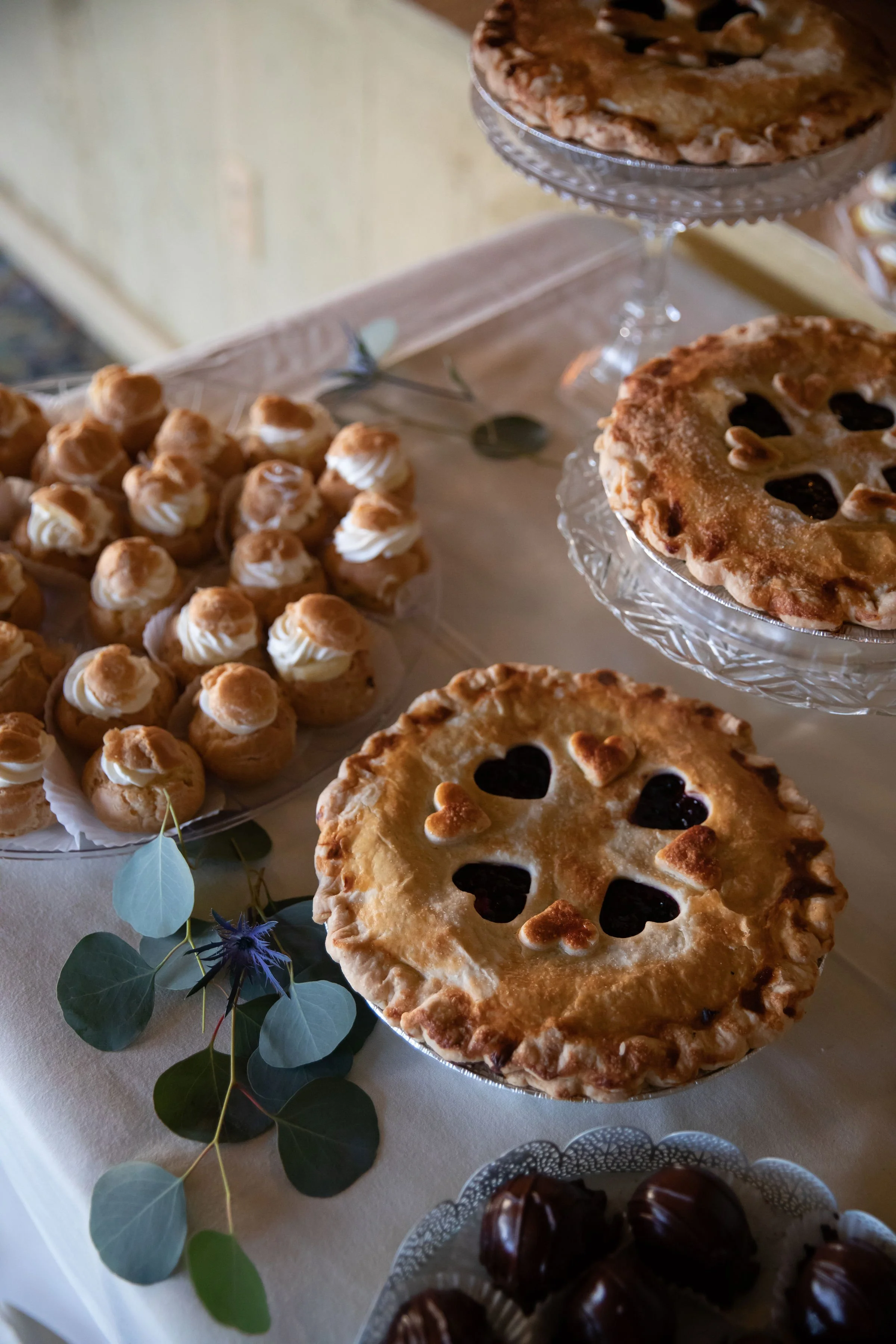 Assorted baked desserts, including heart-shaped lattice pies and cream puffs with meringue topping, displayed on glass and ceramic plates on a white tablecloth.