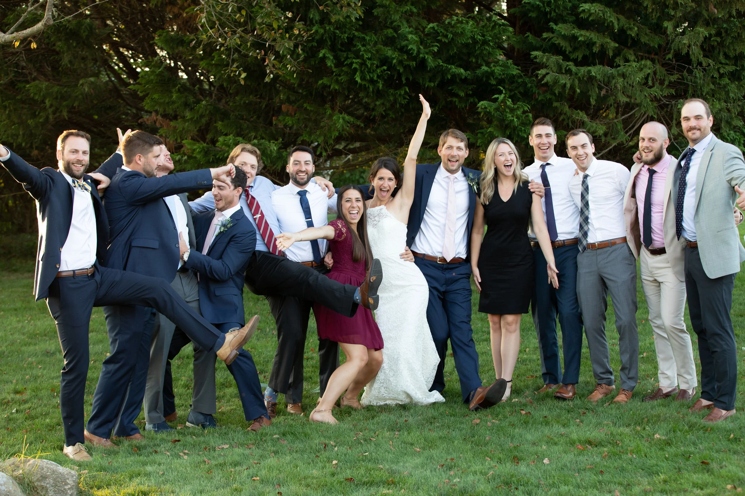 A diverse group of people celebrating outdoors on a grassy field with trees in the background, smiling and laughing, with some playfully lifting a woman in a white dress, indicating a joyful event such as a wedding.
