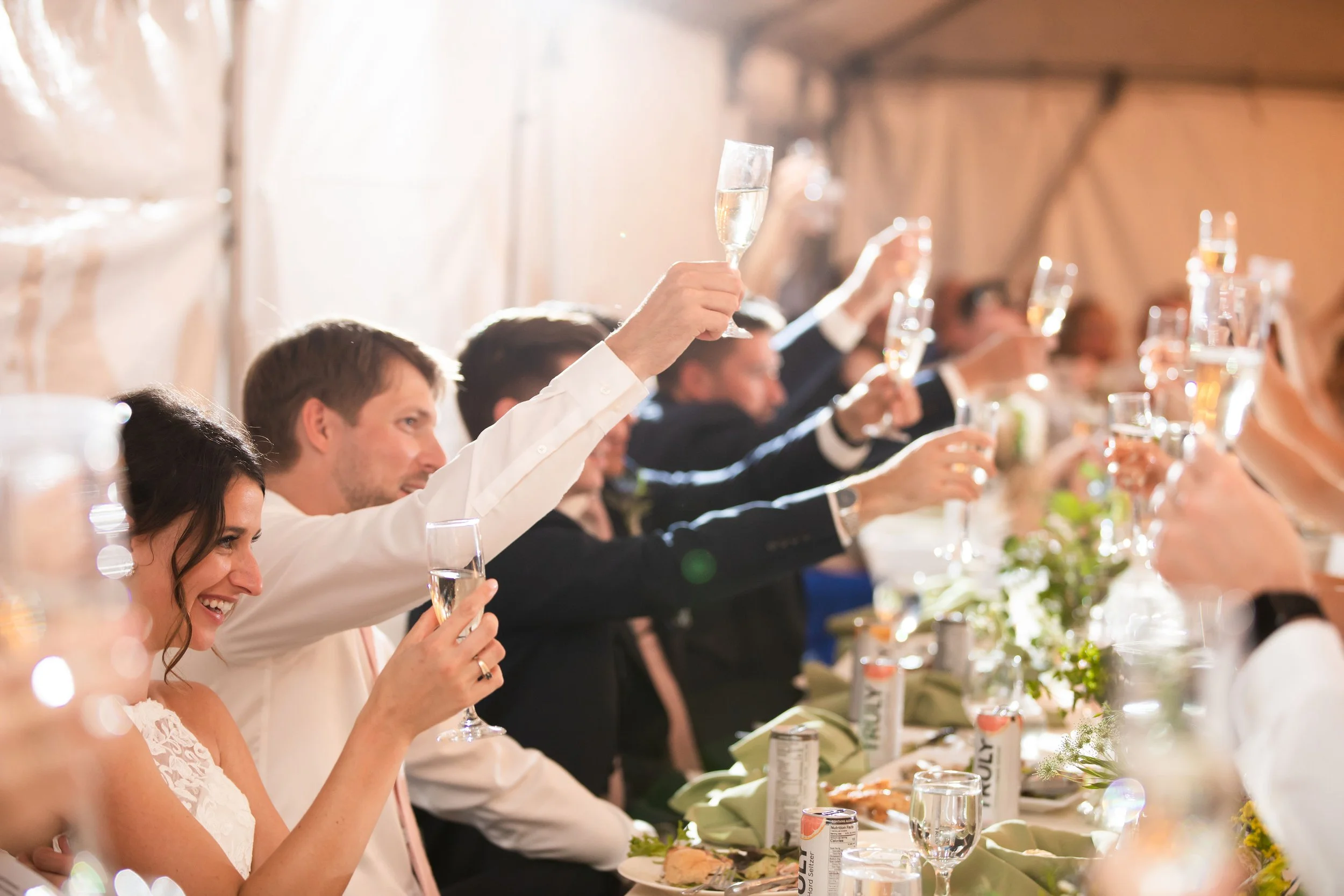People sitting at a wedding reception raising champagne glasses in a toast, with a woman smiling in the foreground.