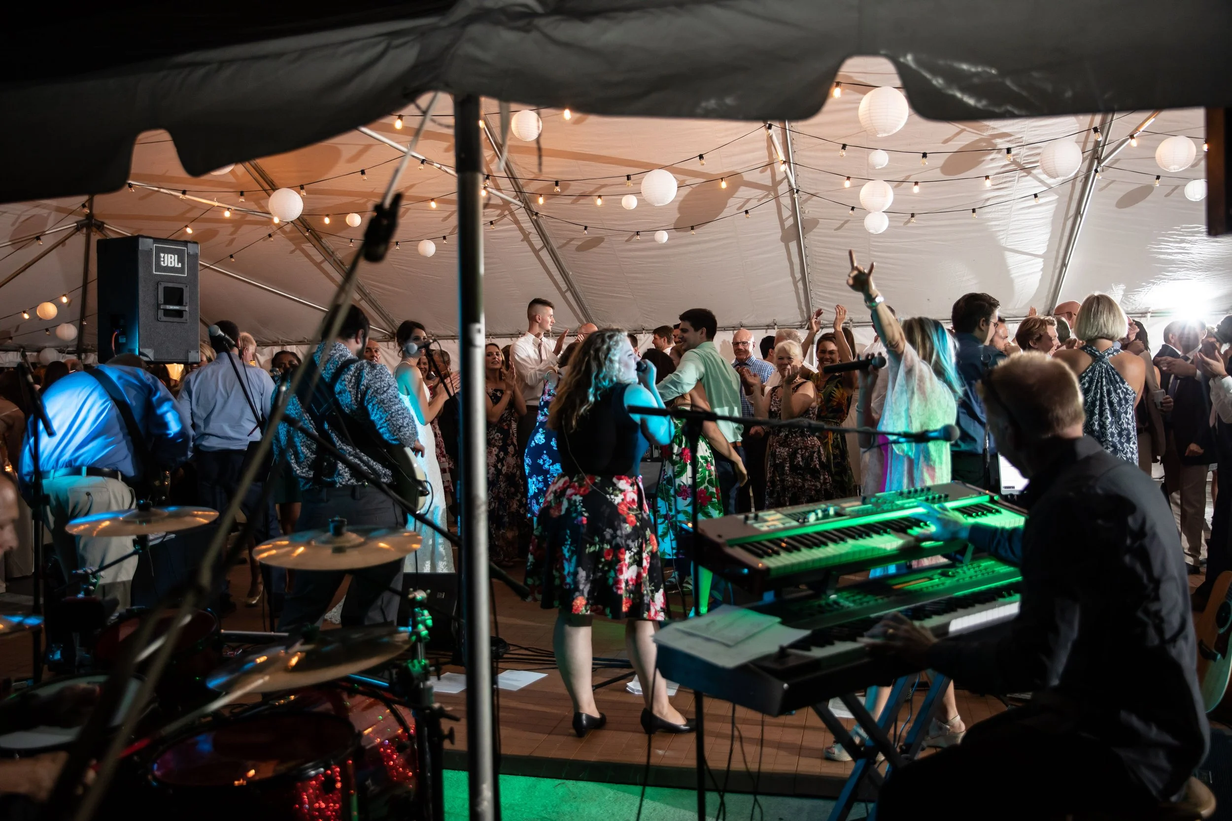 People dancing and enjoying live music under a tent decorated with string lights and paper lanterns, with a pianist and percussion instruments visible in the foreground.