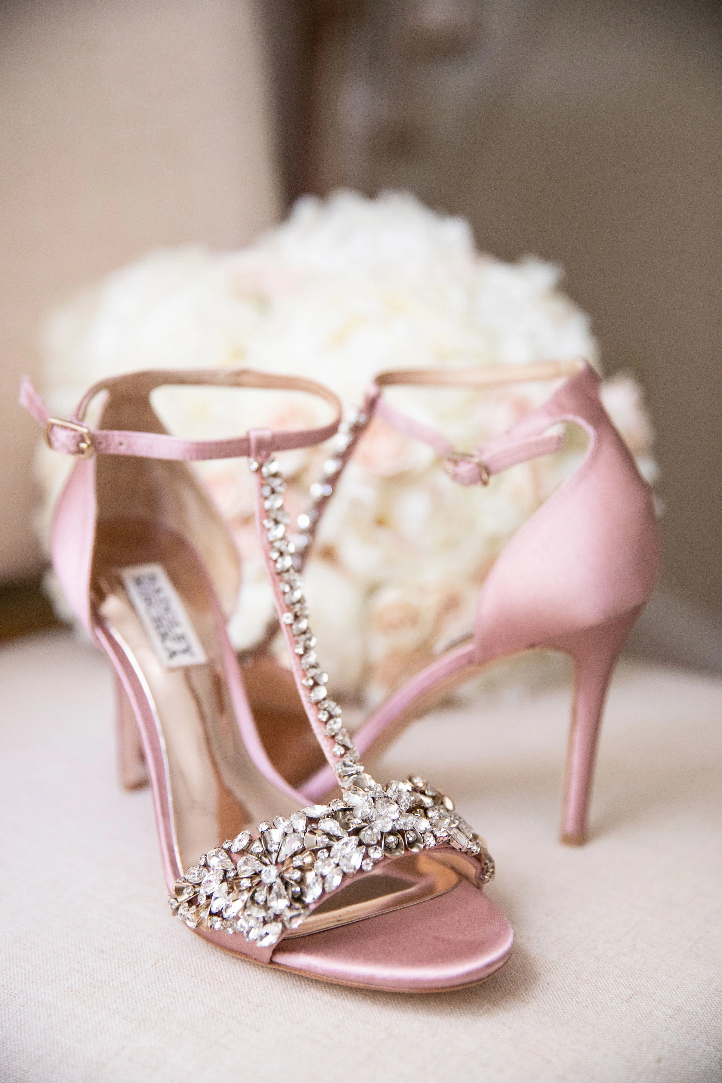 A pair of pink high-heeled shoes with rhinestone embellishments placed in front of a white floral arrangement.