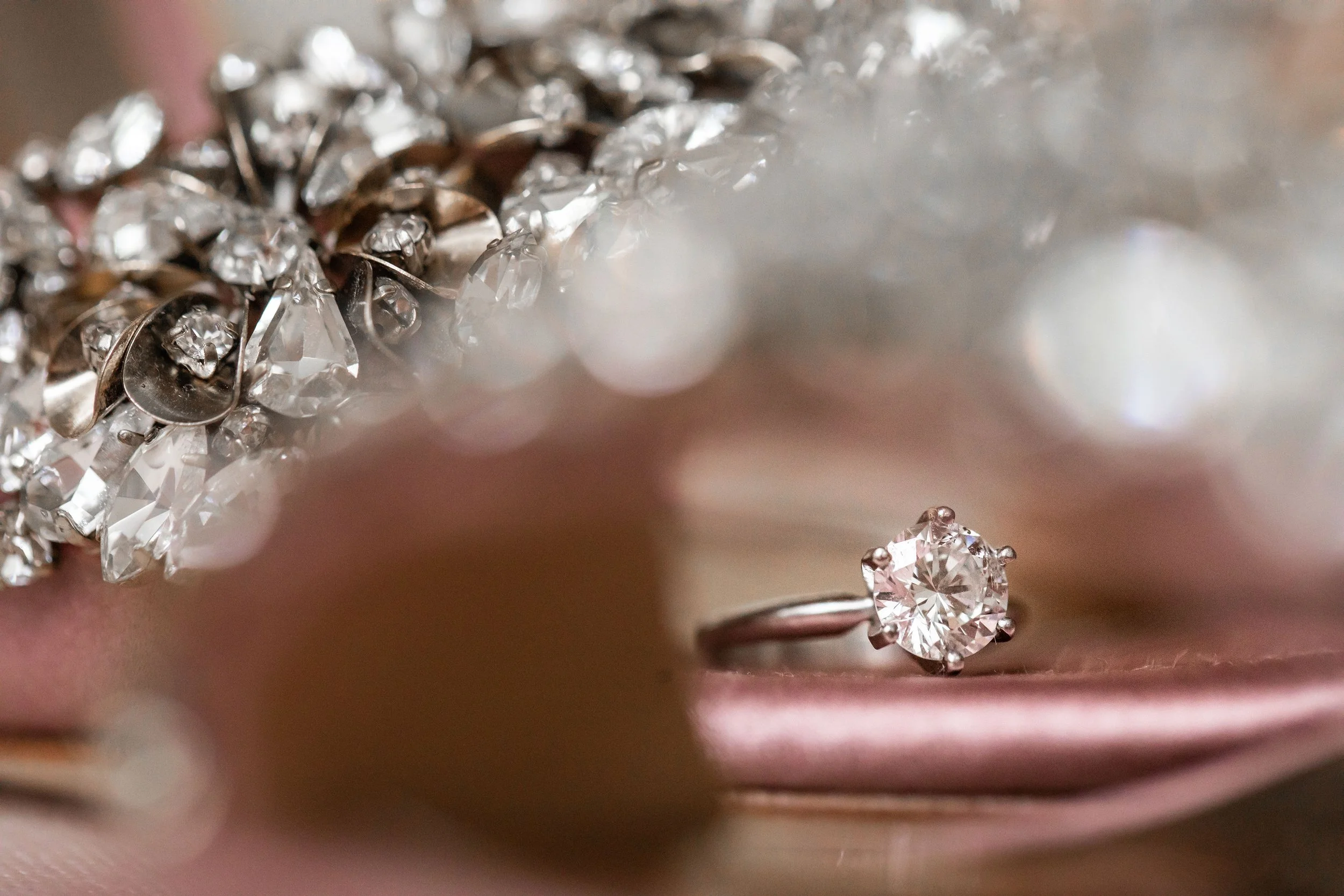 Close-up of a diamond ring on a pink fabric, with a sparkling bracelet or jewelry piece in the background.