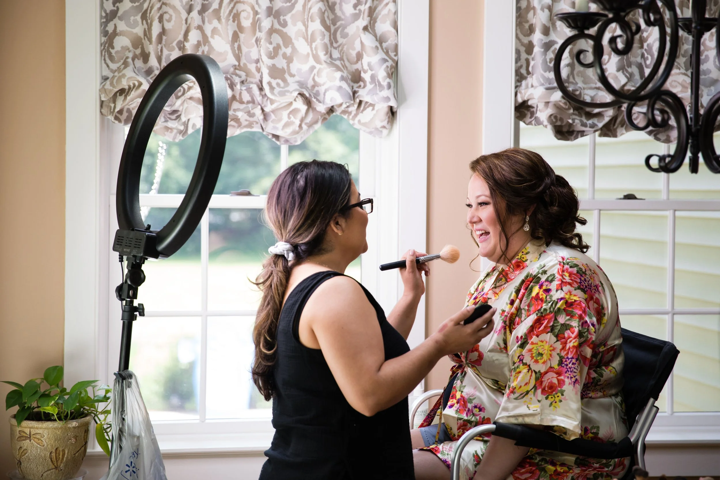 A woman doing makeup on another woman seated in a wheelchair, with a ring light and window in the background.