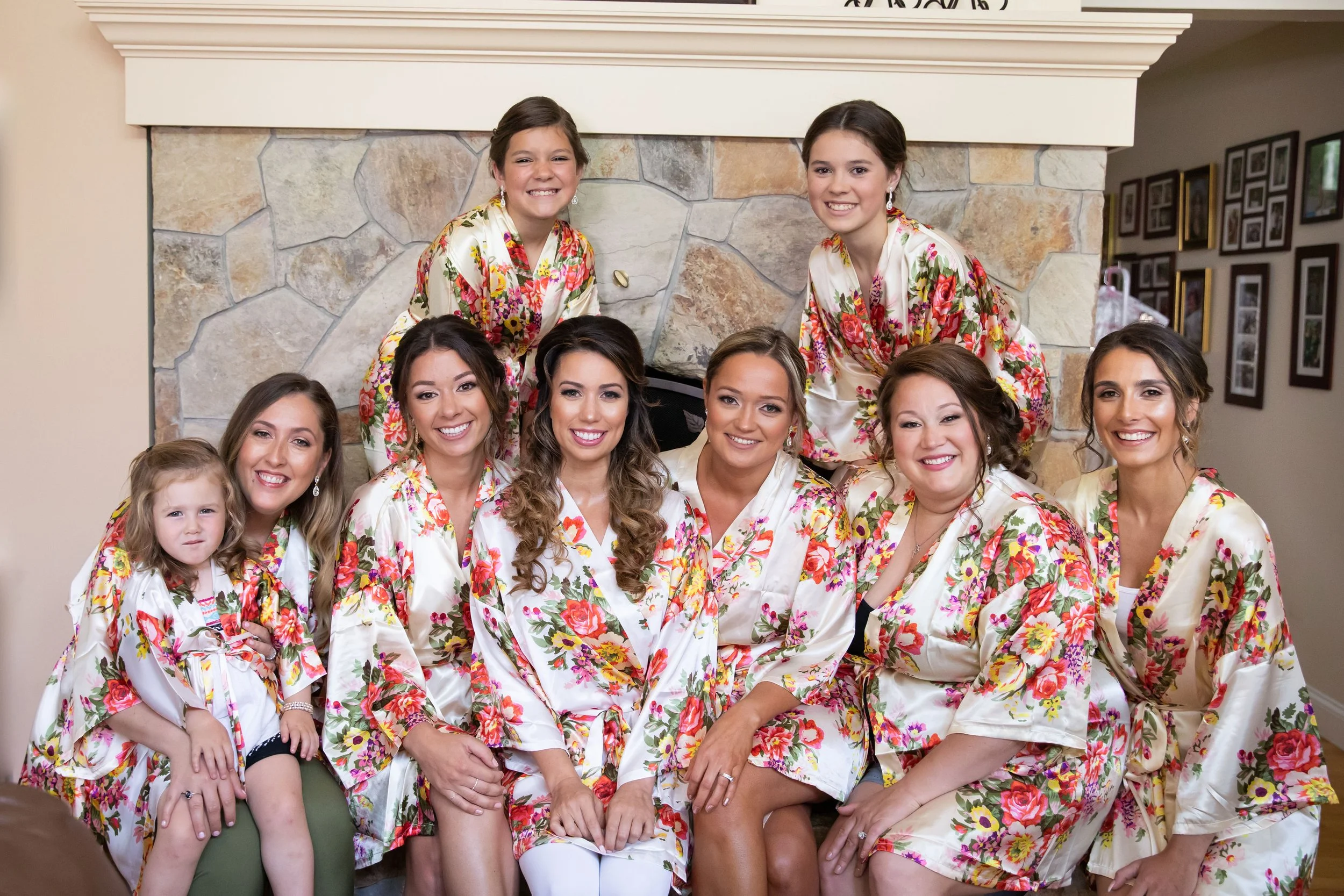 A group of nine women and one young girl smiling and wearing matching floral robes, gathered in front of a stone fireplace for a bridal party photo.